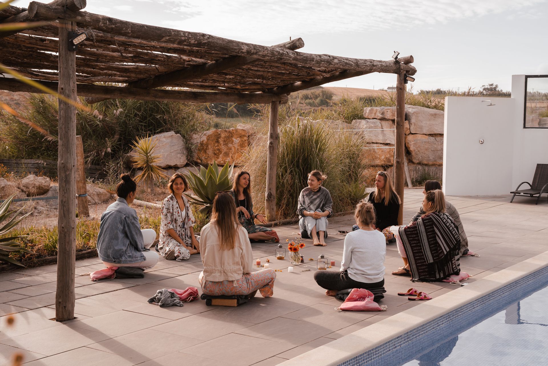 A group of people sit in a circle under a wooden pergola near a pool, engaged in an activity.