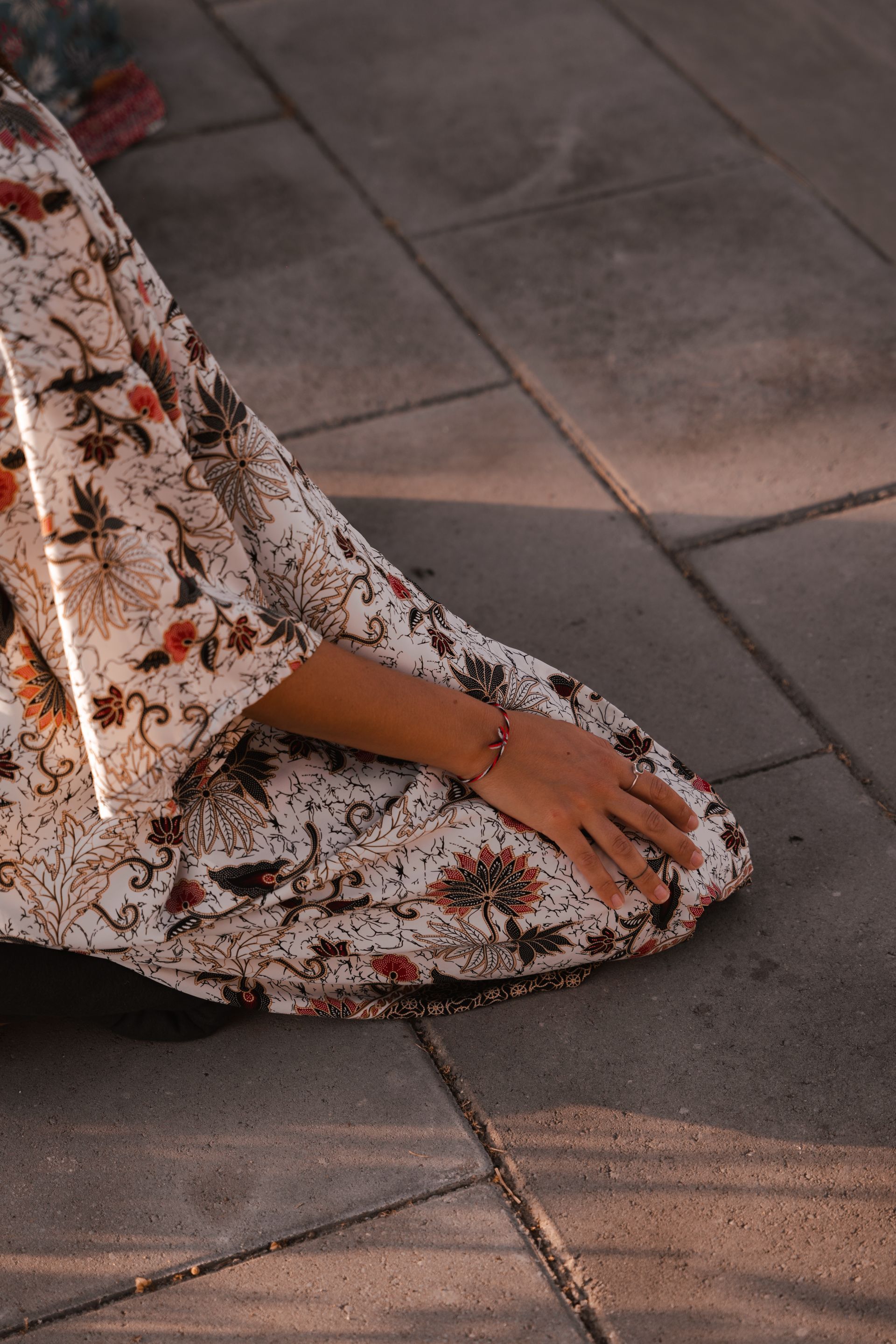 Person kneeling on paved ground, wearing a floral-patterned dress, arm resting with bracelet.