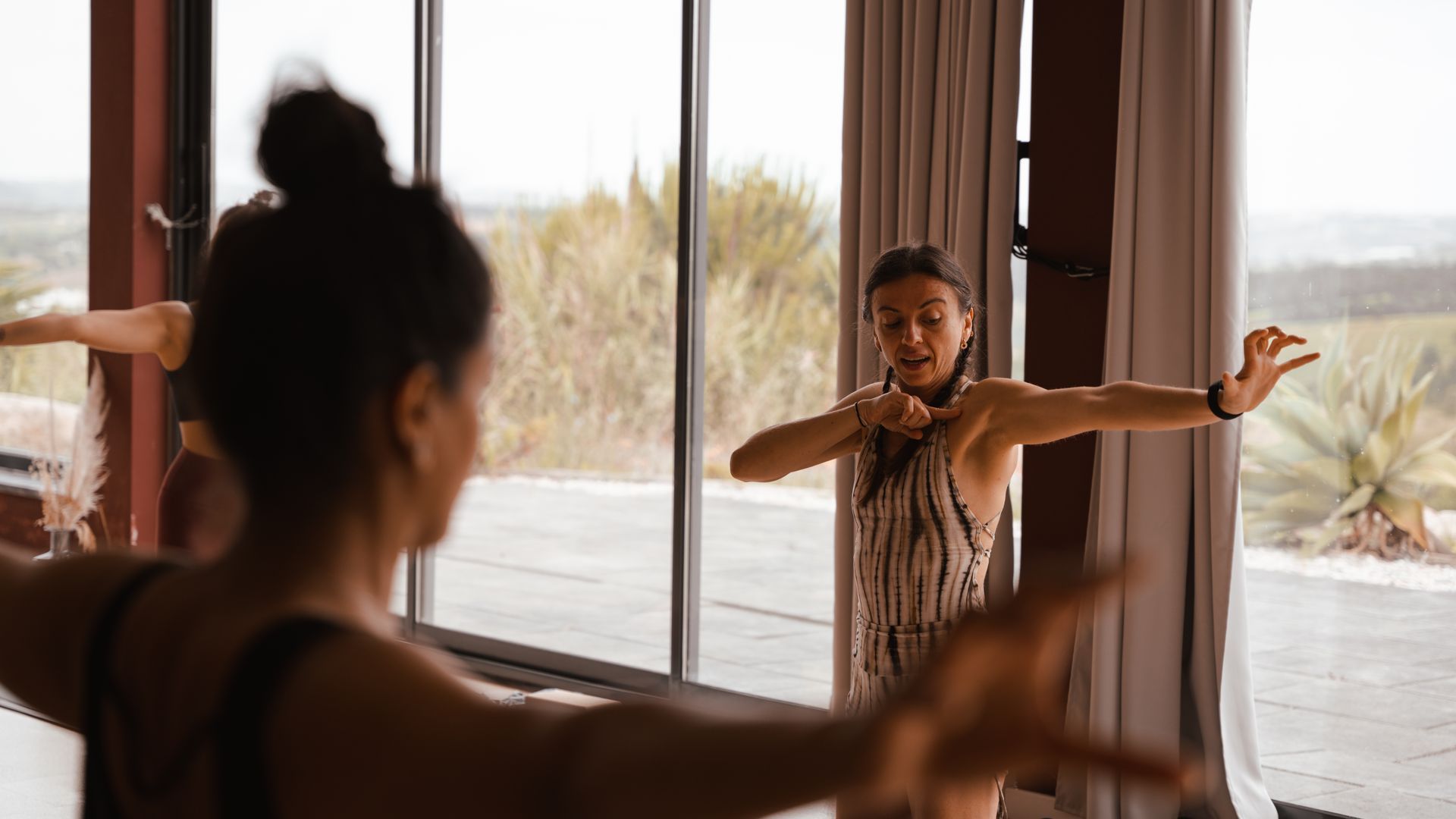 People in a dance studio with instructor, arms extended near windows.