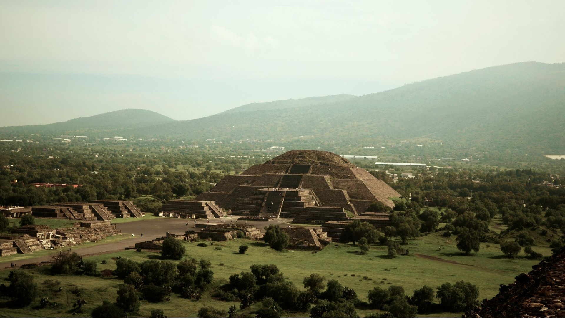 Pyramid of the Sun in Teotihuacan, Mexico, with surrounding buildings and green landscape against a mountain backdrop.