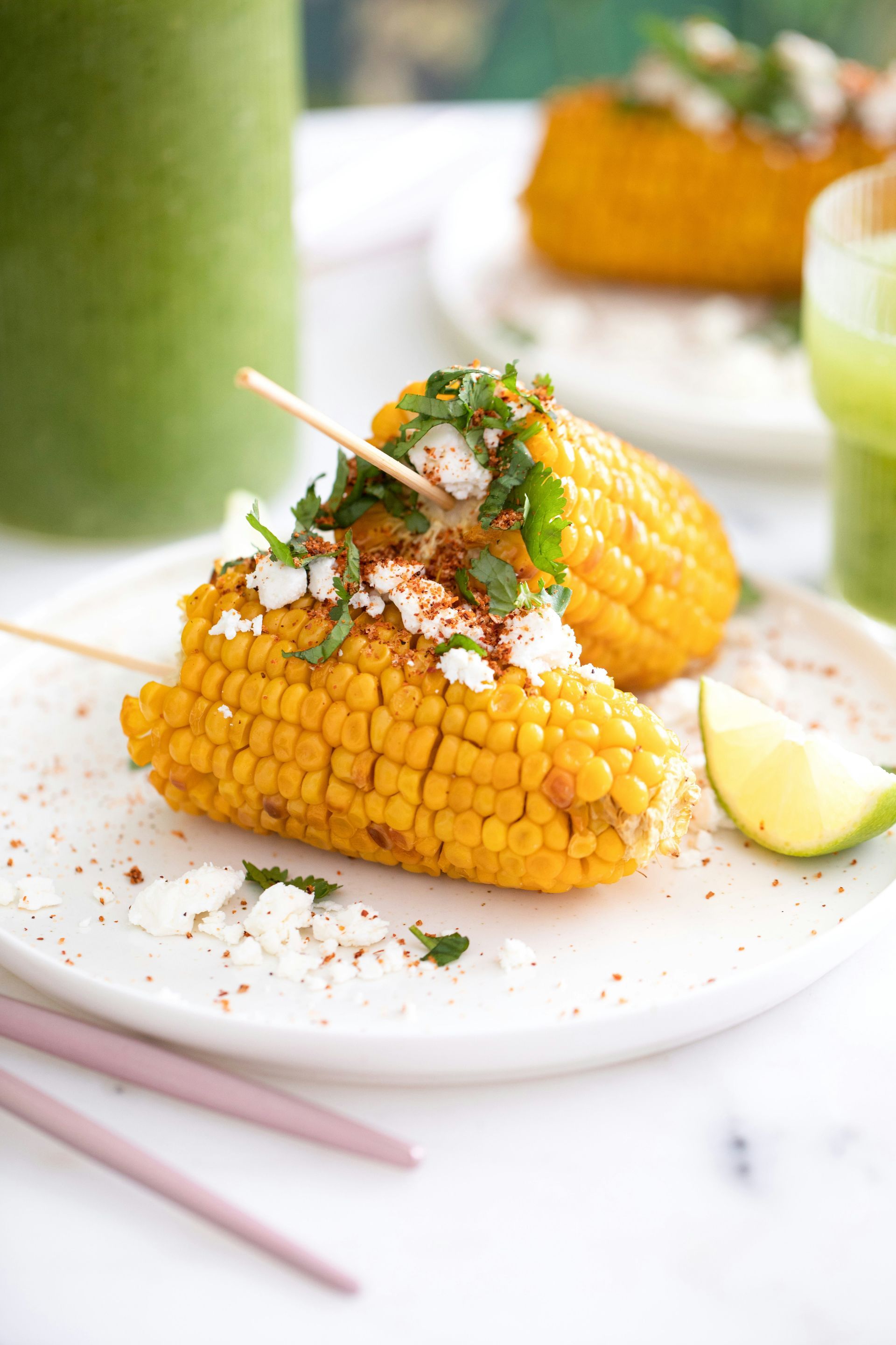 Grilled corn on the cob with toppings, served on a white plate with a lime wedge and a green drink.