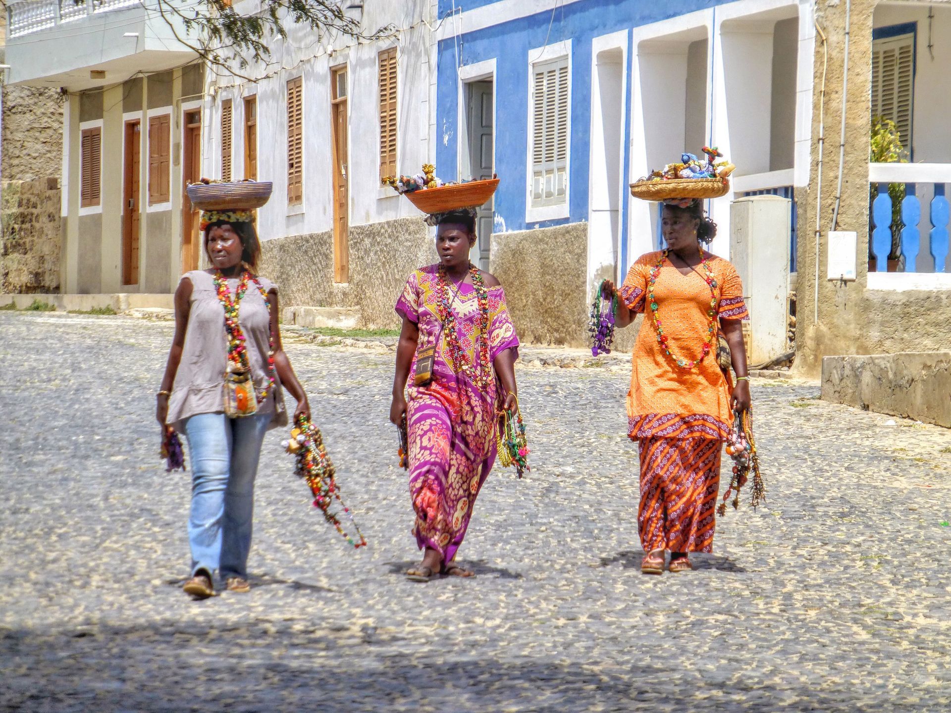 Three women walking on cobblestone street, each carrying baskets on their heads and colorful items. Buildings in background.