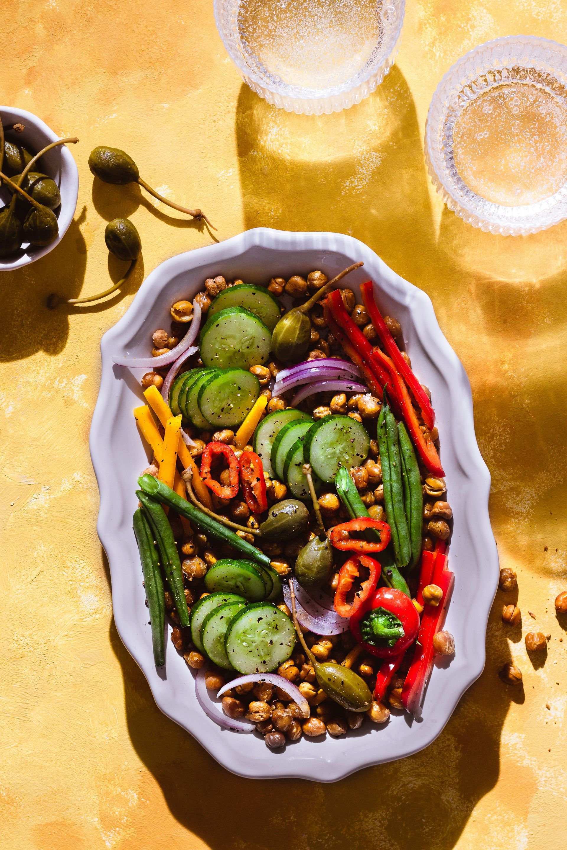 Colorful chickpea salad with cucumbers, peppers, red onion, capers, and two glasses of white wine.