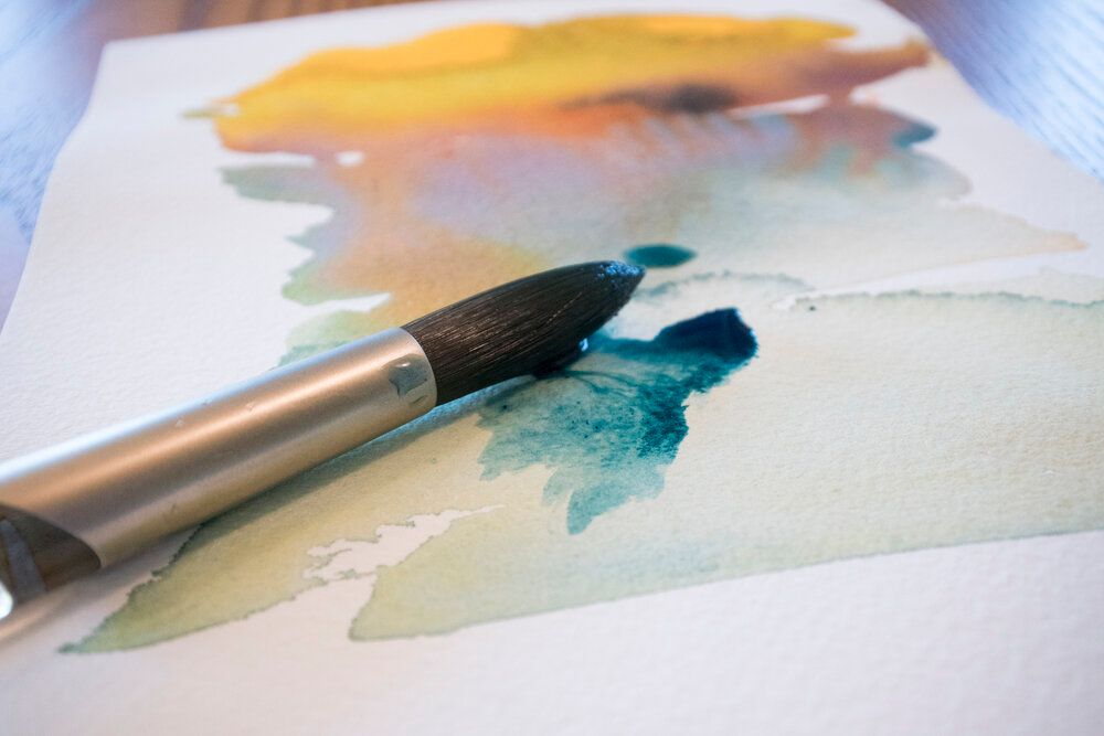 Watercolor brush adding teal paint to paper with yellow and orange washes.