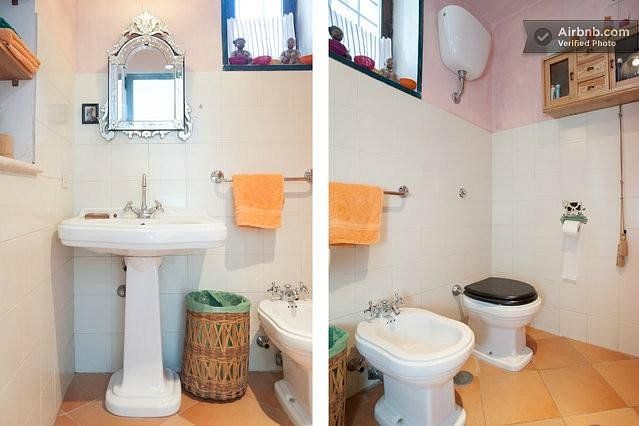 Bathroom with a pedestal sink, mirror, bidet, toilet, and orange towels. Beige walls, window, and wicker wastebasket.