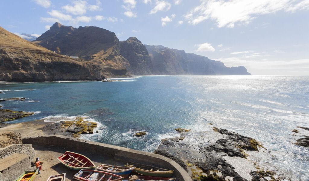 Coastal view with mountains, ocean, and boats on the shore under a blue sky with clouds.