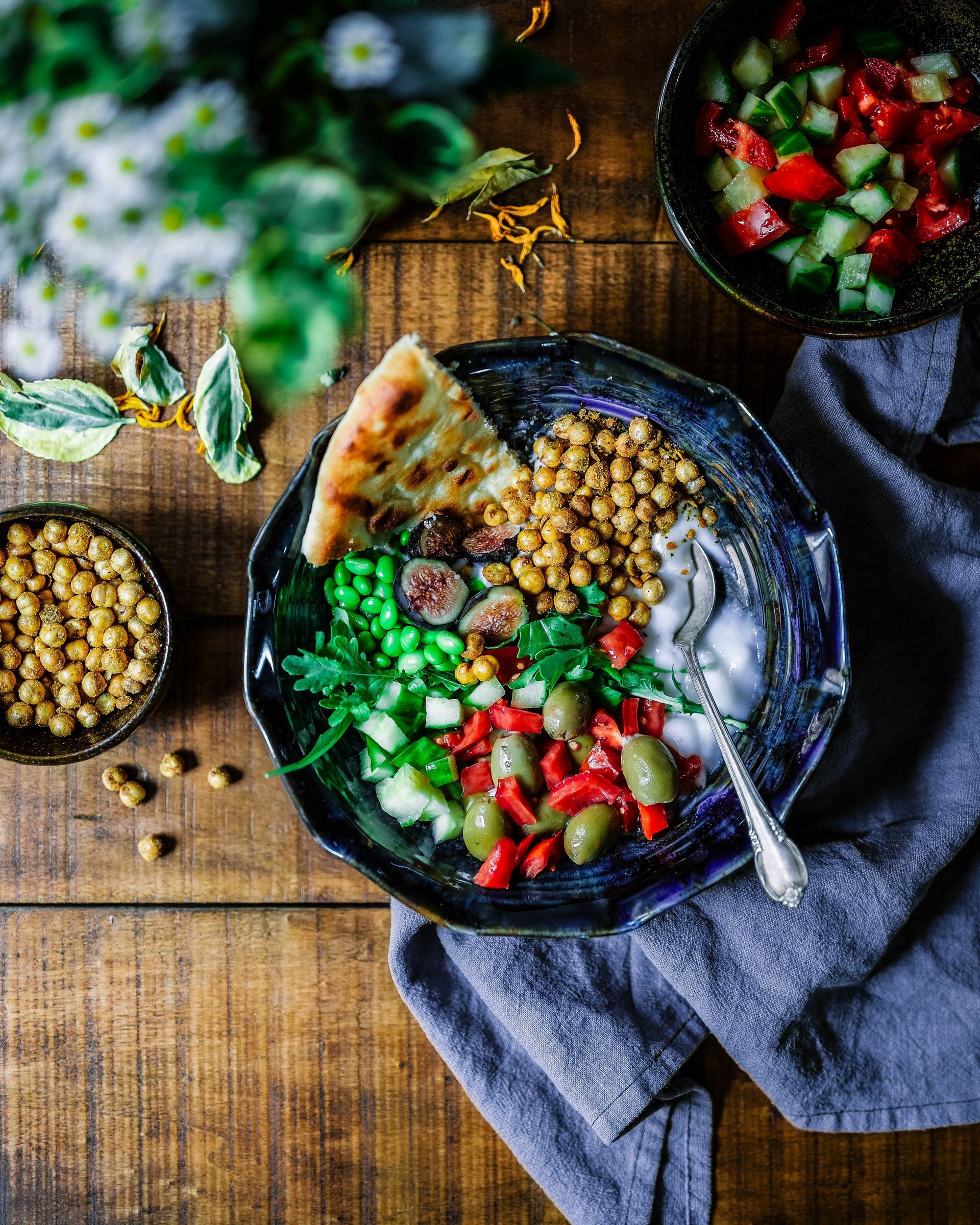 Colorful bowl of food on a wooden table with chickpeas, olives, pita bread, and a small bowl of salad.