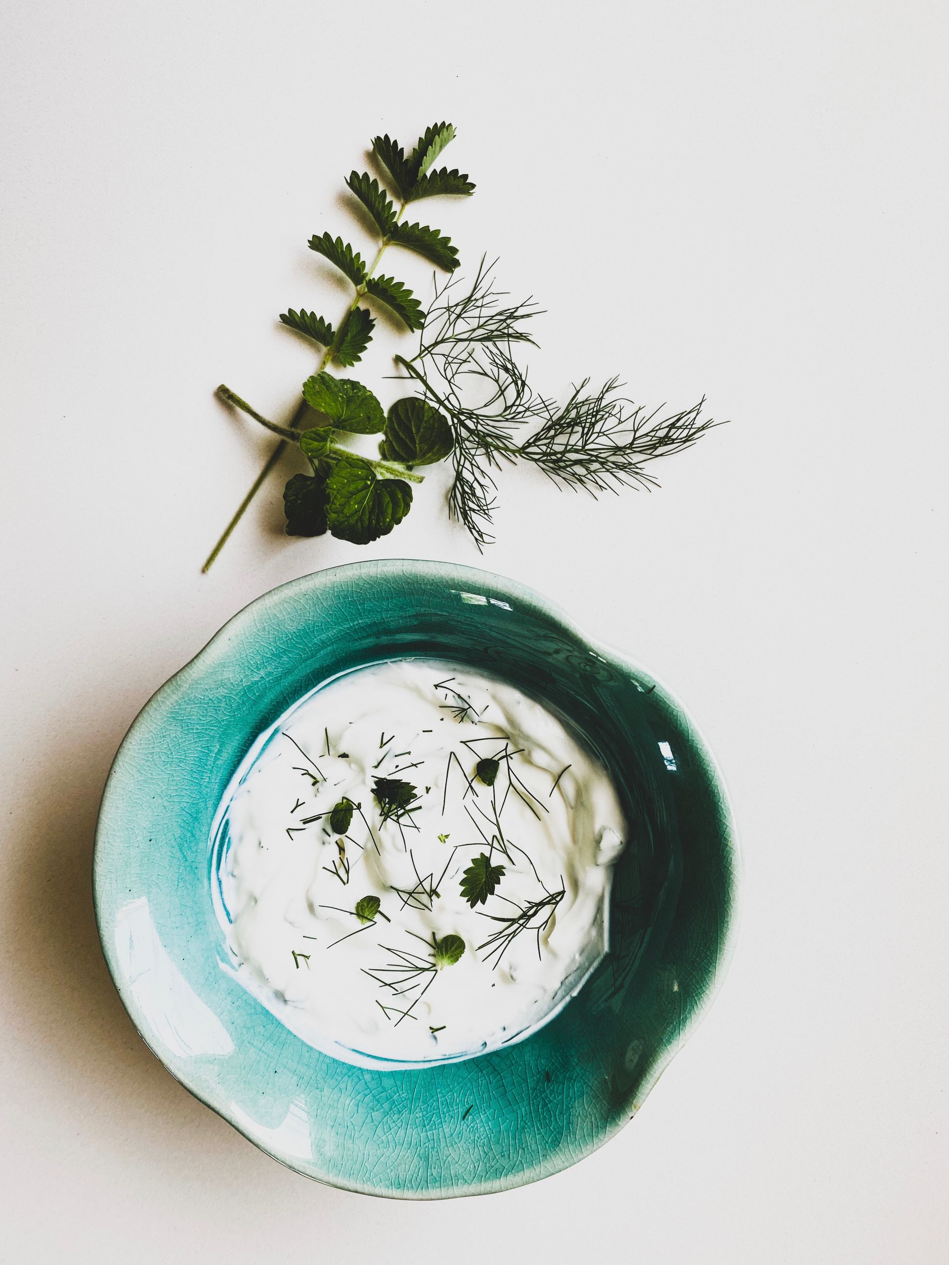 White dip in teal bowl garnished with herbs, on a white surface.