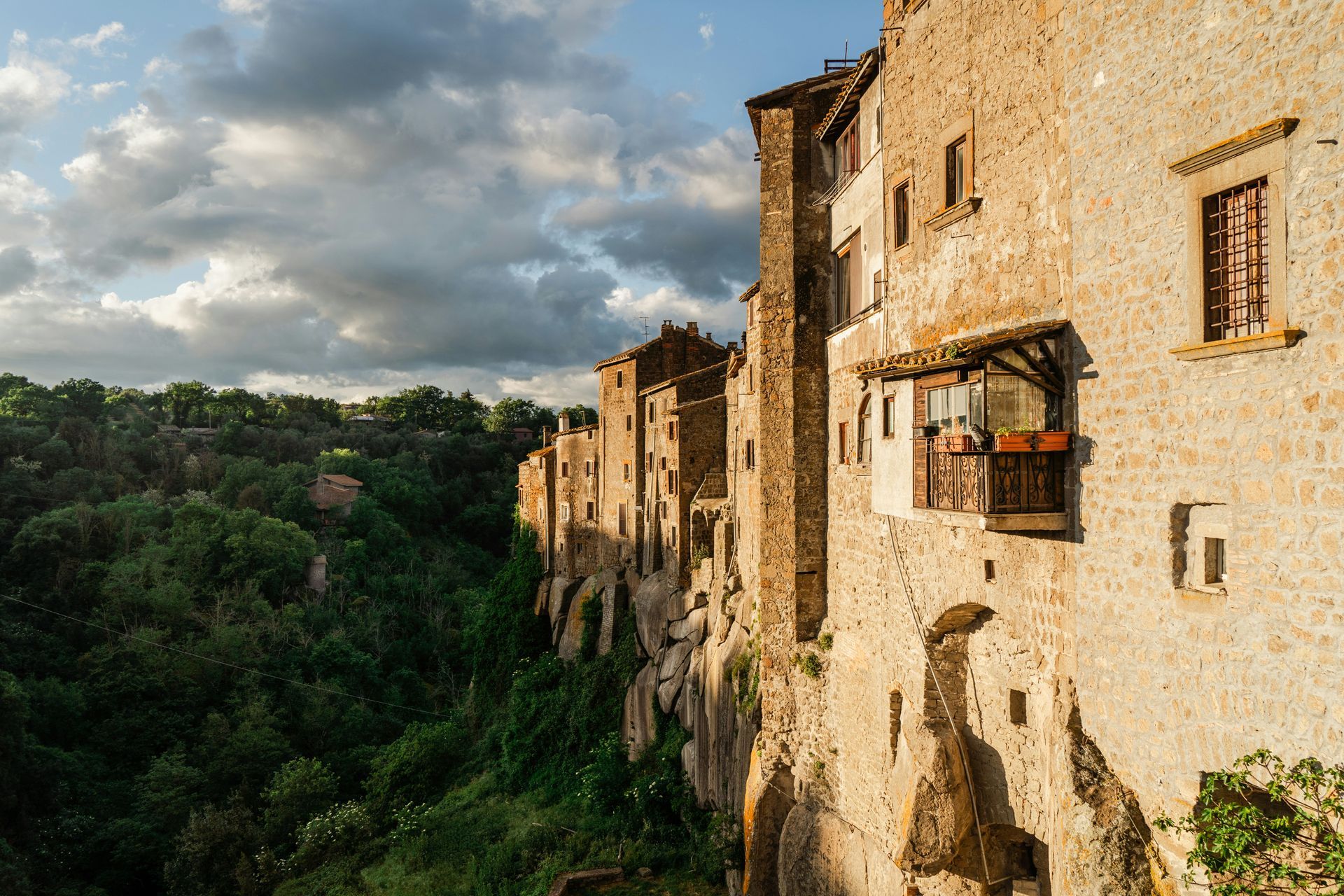 Cliffside buildings, bathed in sunlight, overlook a lush green valley with a cloudy sky.