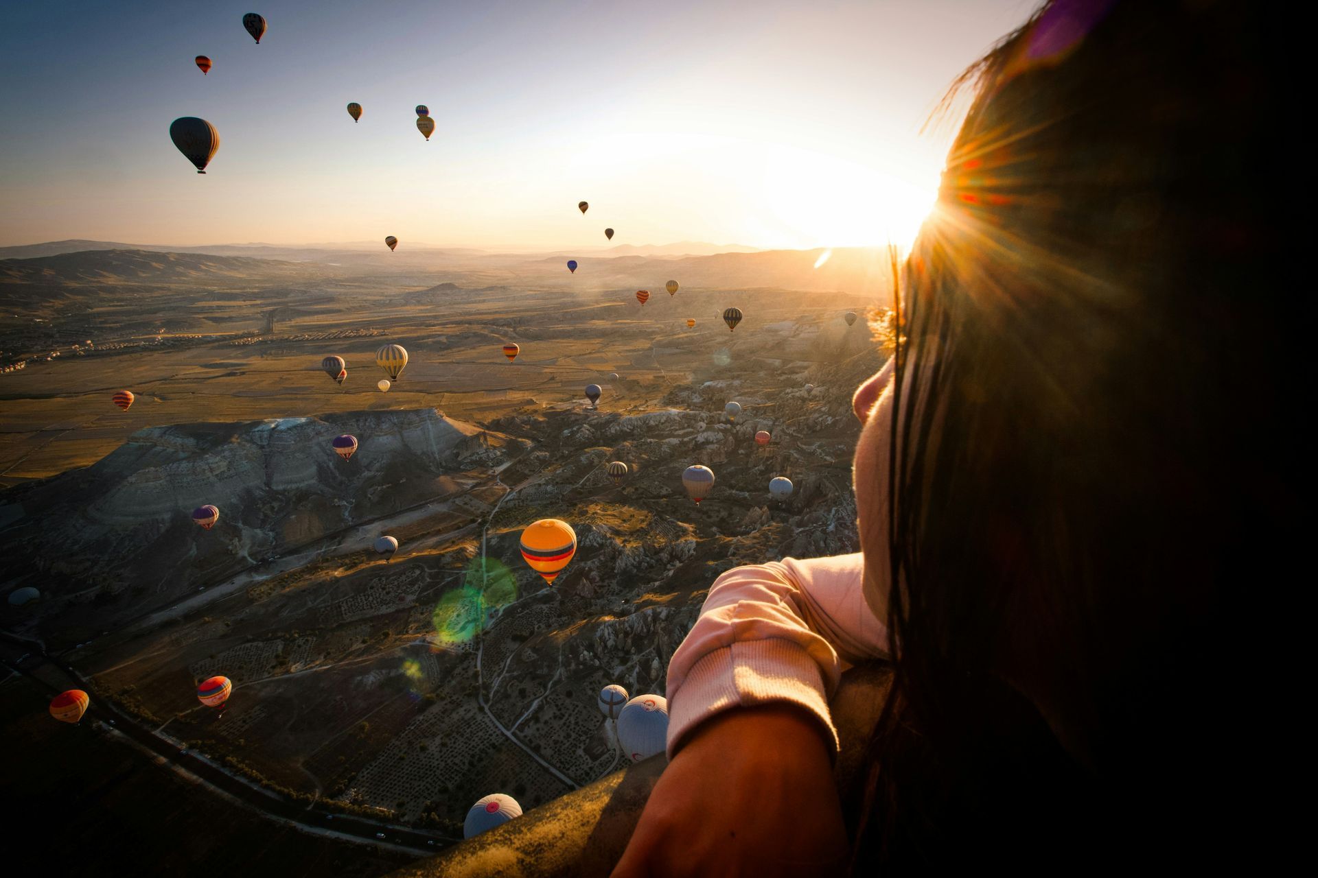 Woman watches hot air balloons over a valley at sunrise. Bright sun, colorful balloons.