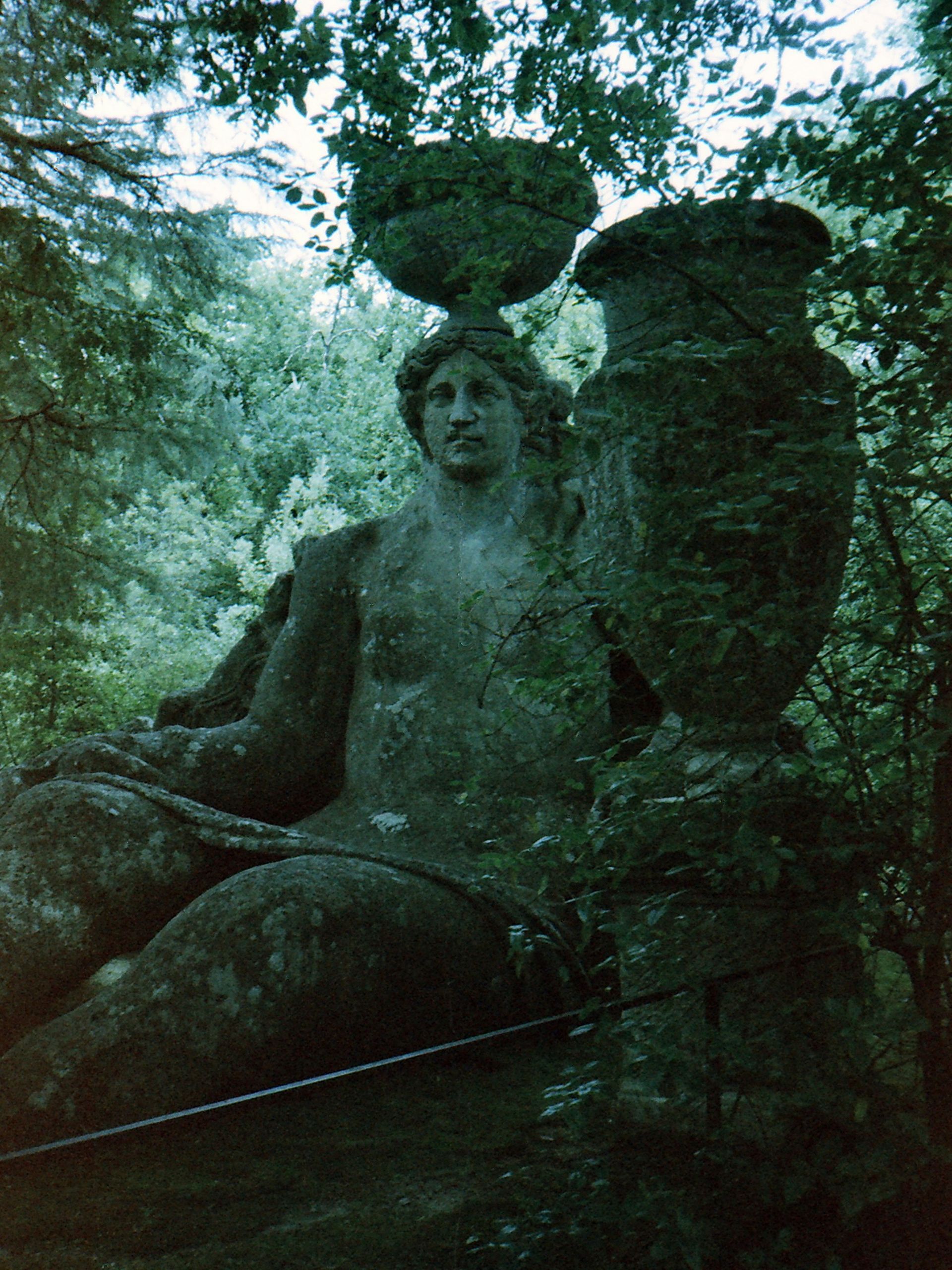 Stone statue of a seated figure holding a vessel and a bowl, surrounded by foliage.