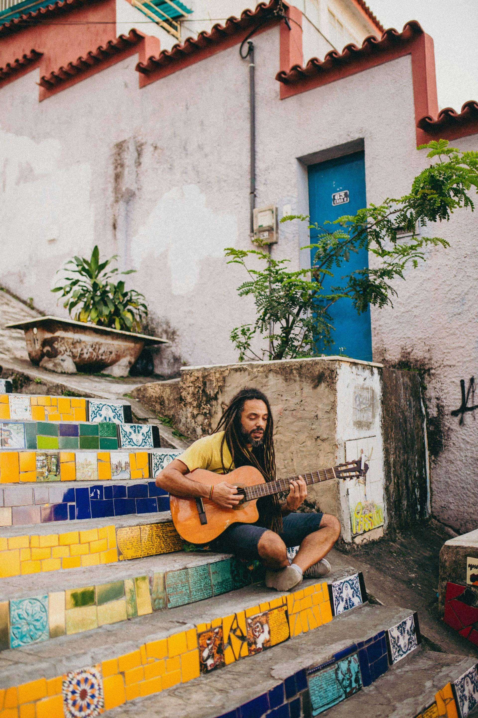 Man playing guitar on colorful mosaic stairs in front of a white building with a blue door.