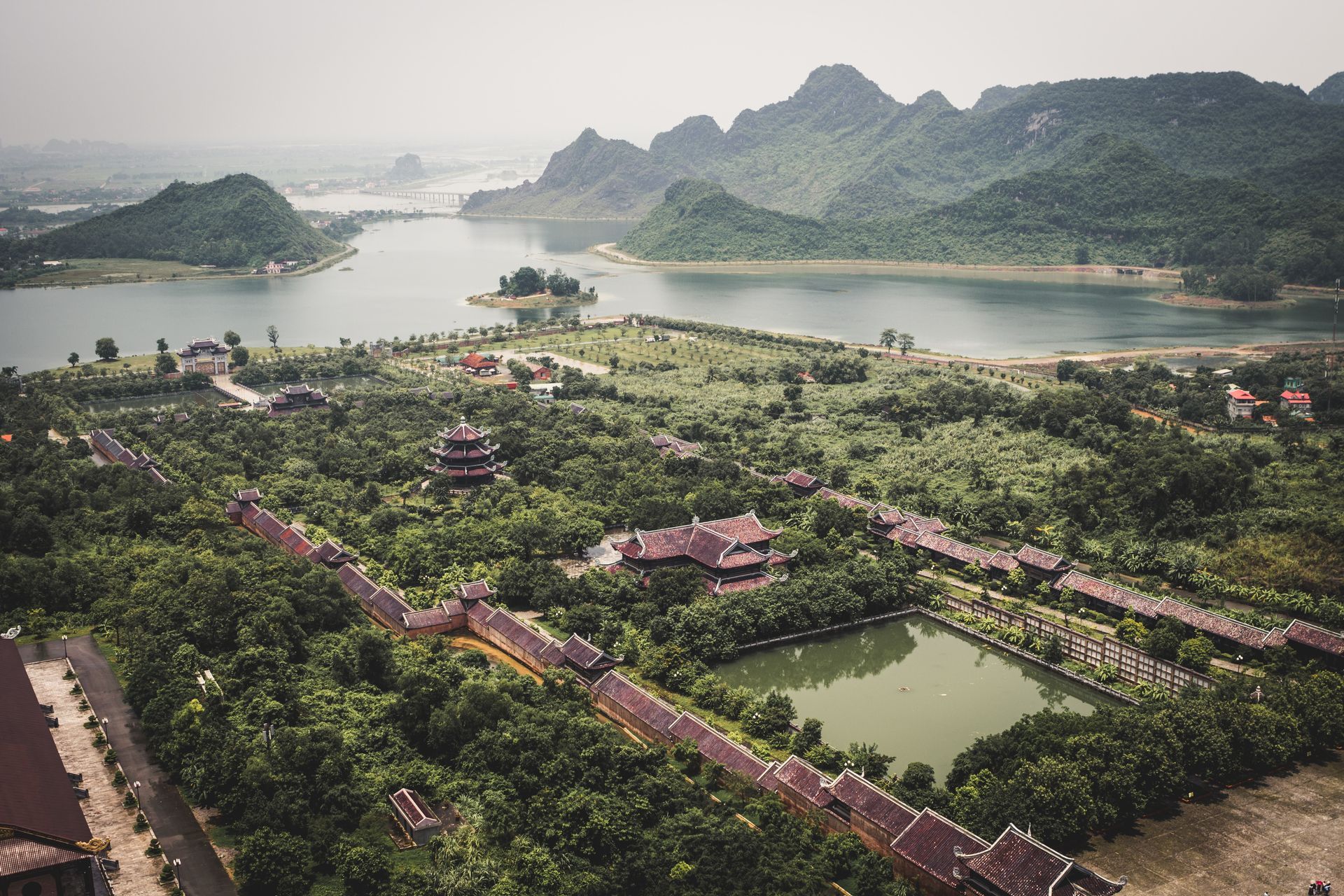 Aerial view: lush temple complex with lake and mountains in background.
