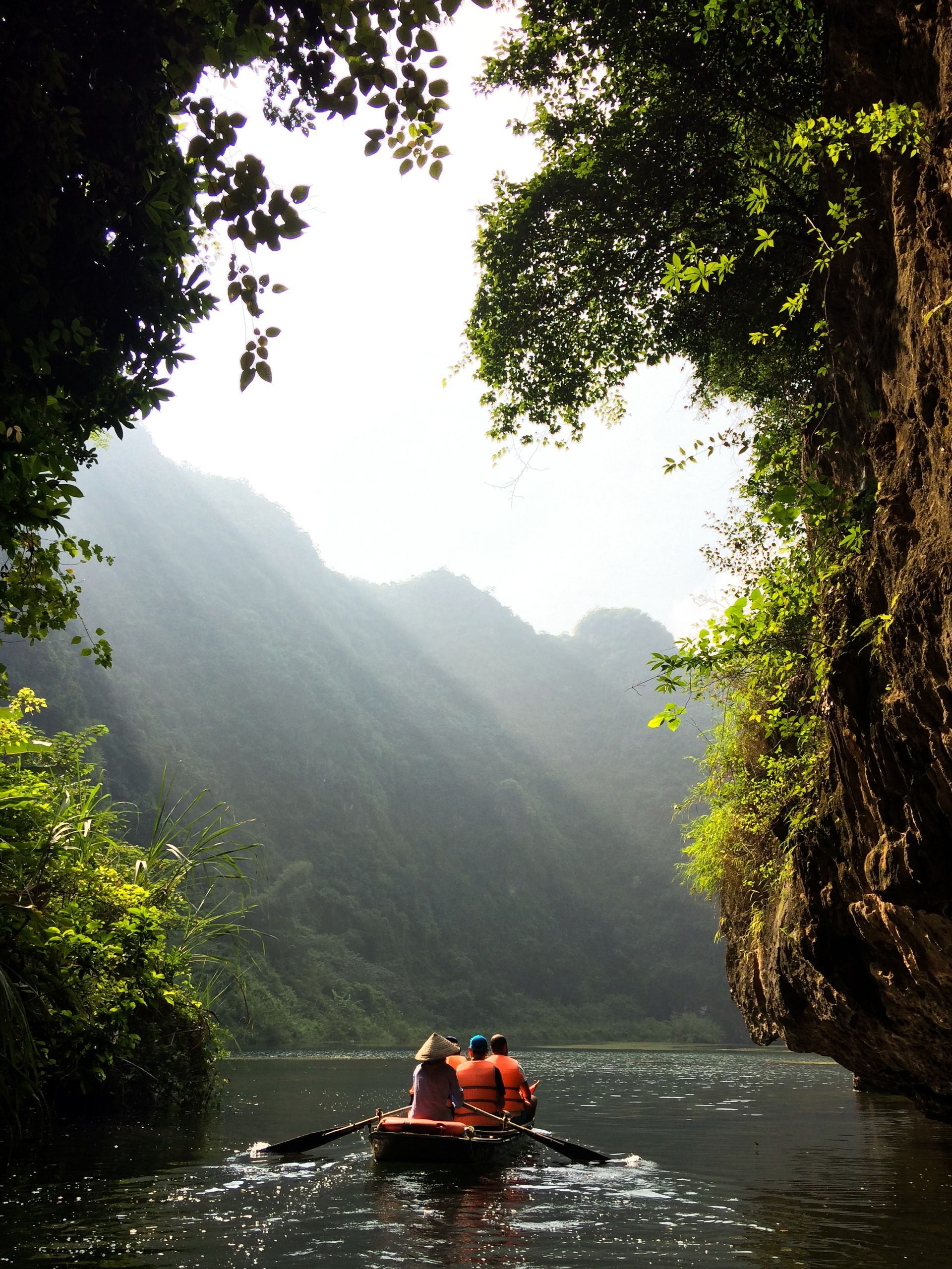 Boat with passengers on a river, framed by lush green trees and cliffs, with mountains in the background.