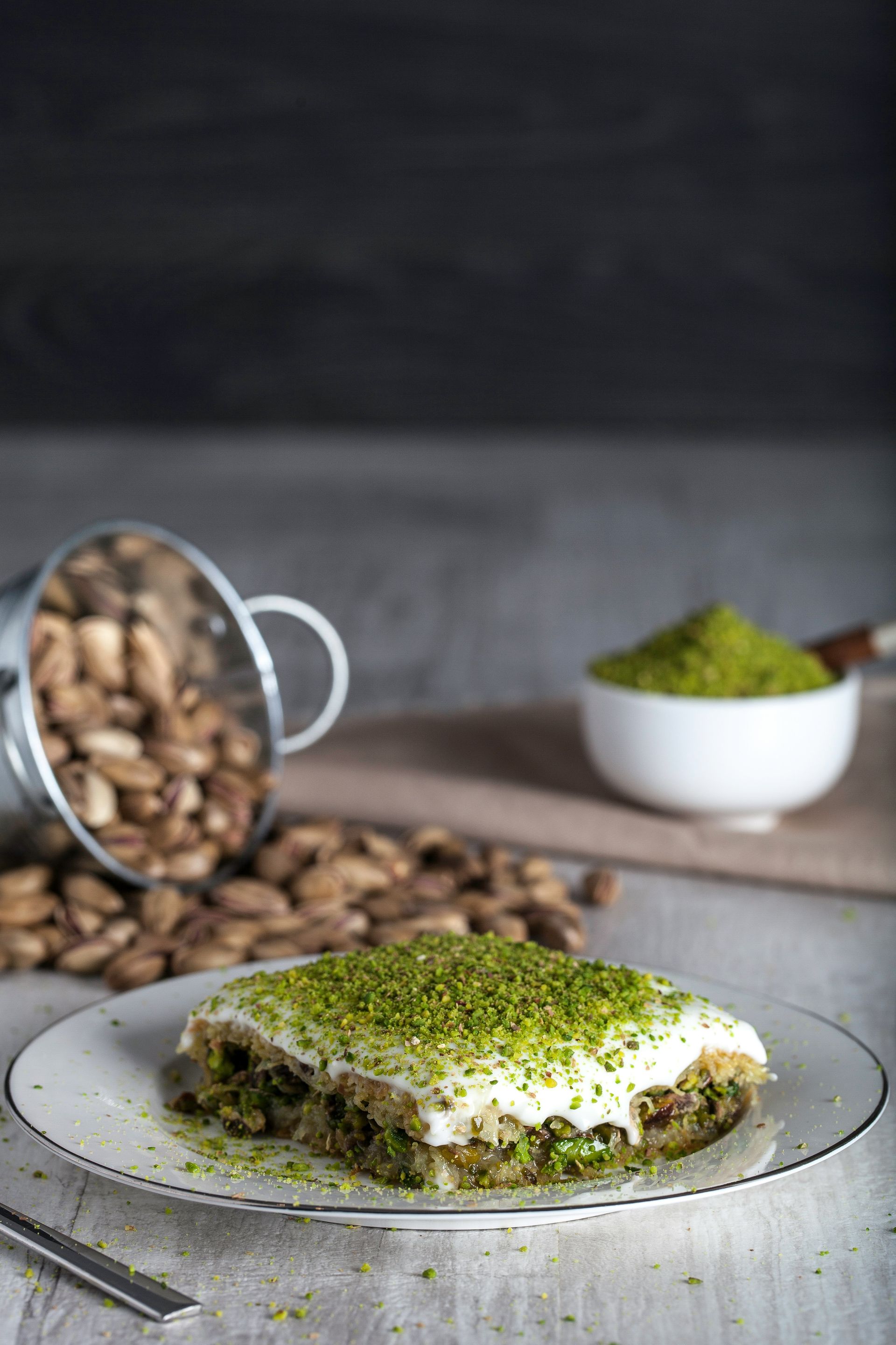 Baklava dessert topped with pistachio, served on a white plate with nuts nearby and a small bowl of ground pistachio.