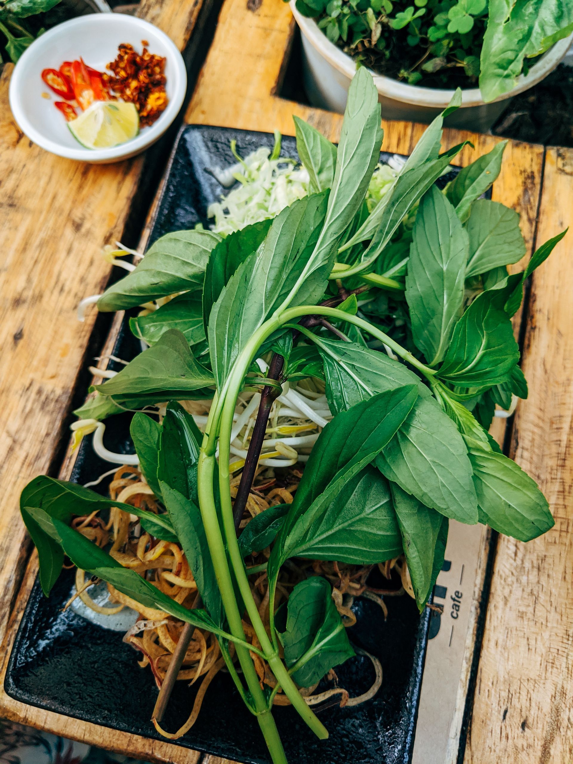 Fresh herbs and vegetables on a black plate, with chili paste and lime, ready for a Vietnamese dish.