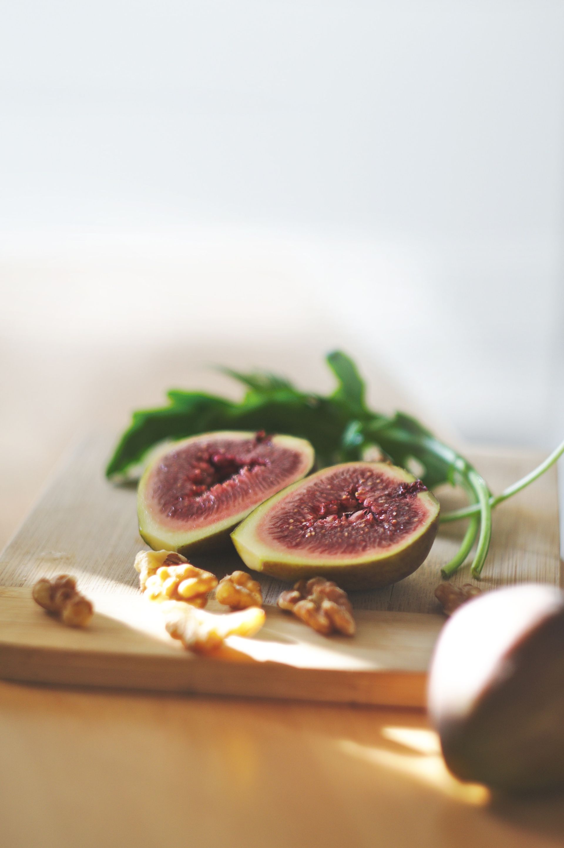 Half a fig, walnuts, and greens on a wooden cutting board, with another fig out of focus.