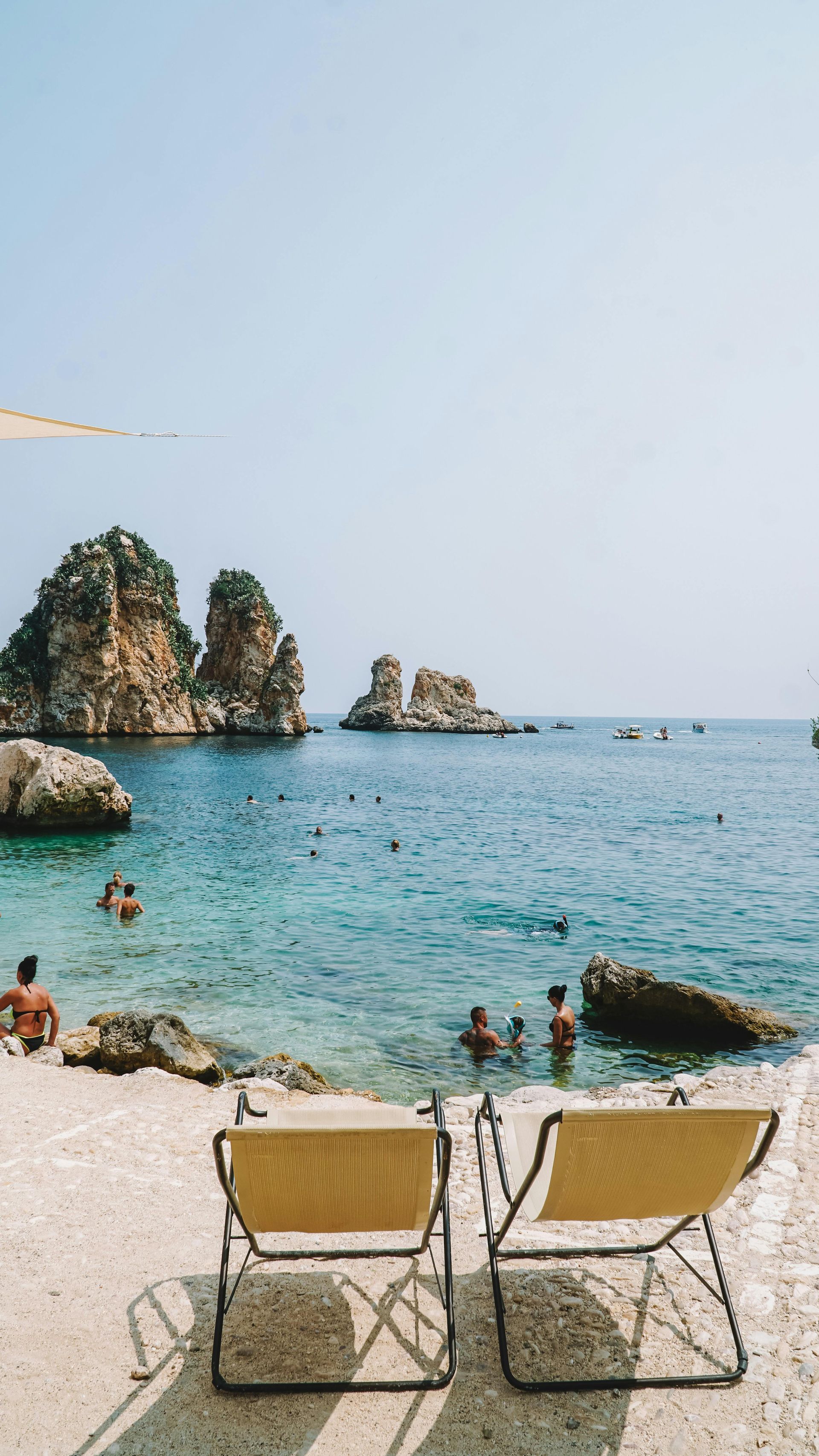 Two beach chairs on a sandy beach looking out at rocky cliffs in turquoise water. People swim.