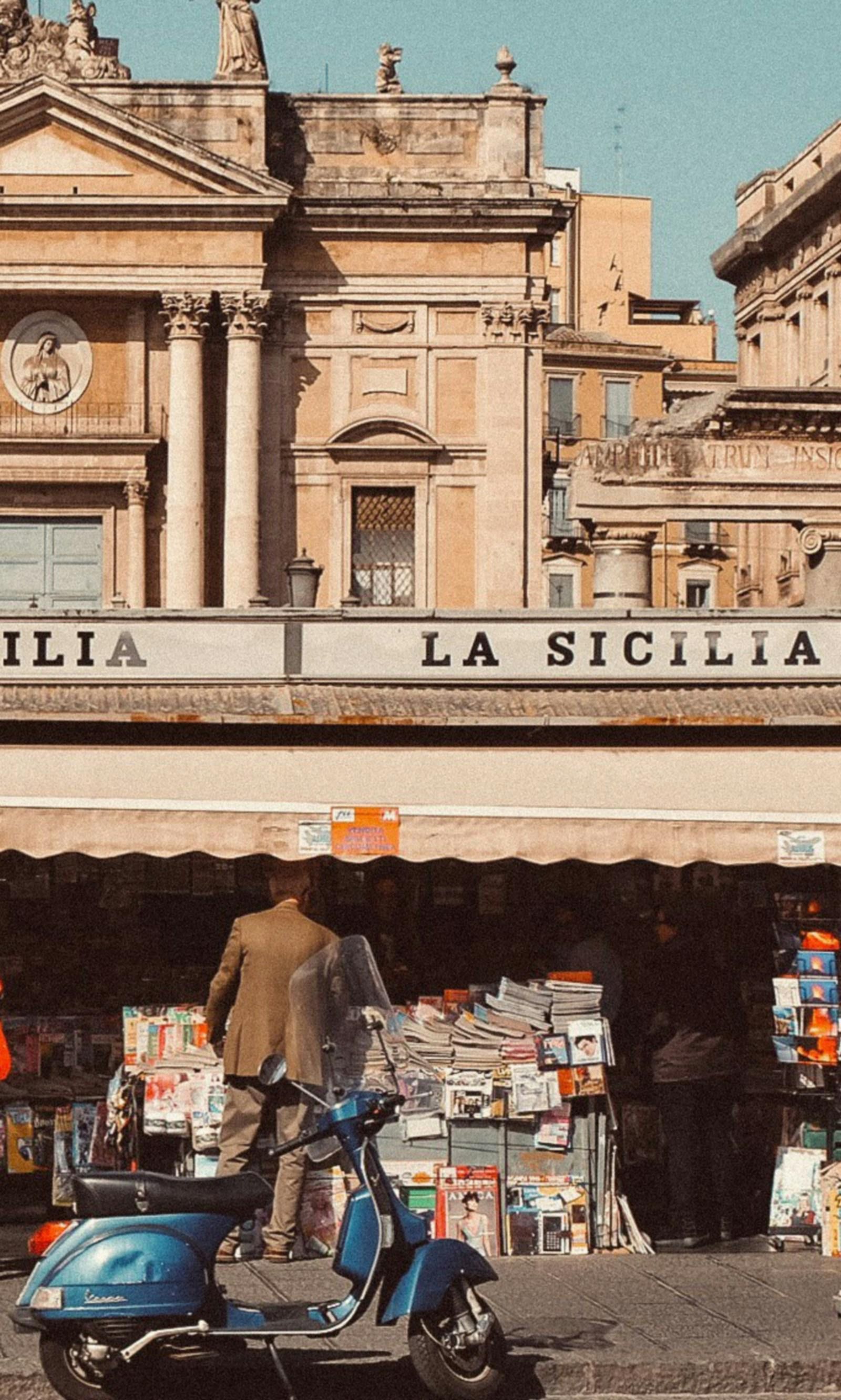 A man standing near a blue scooter in front of a newsstand.