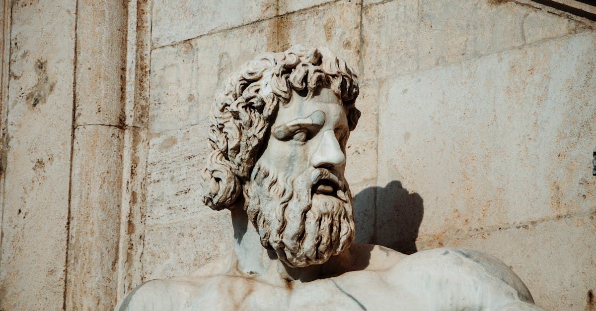 Statue of a bearded man with curly hair and stern expression, marble against a light-colored wall.