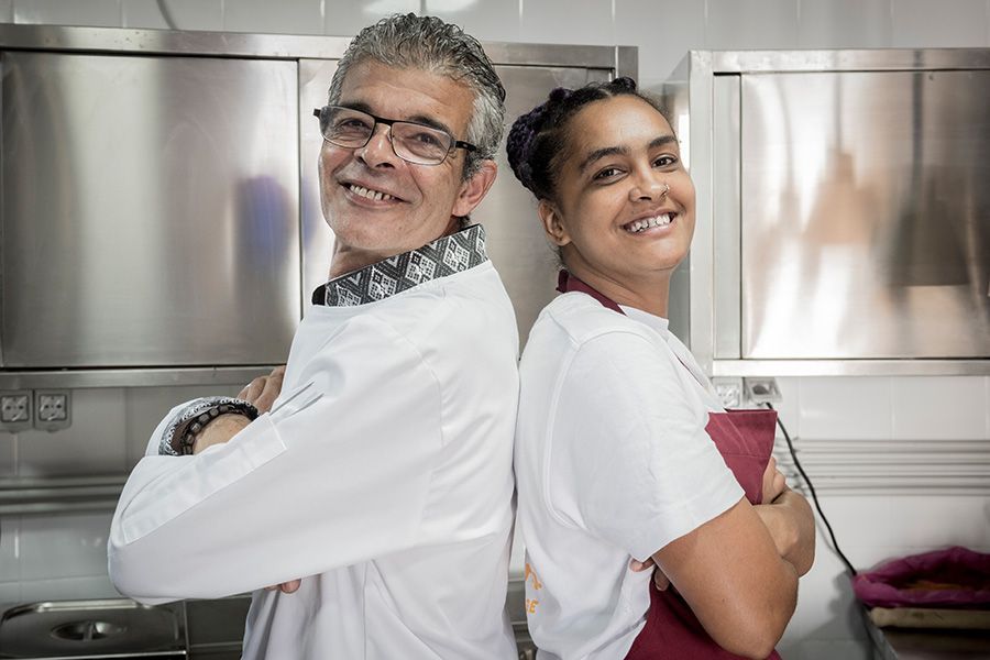 Two people in a kitchen smile, standing back-to-back with arms crossed.