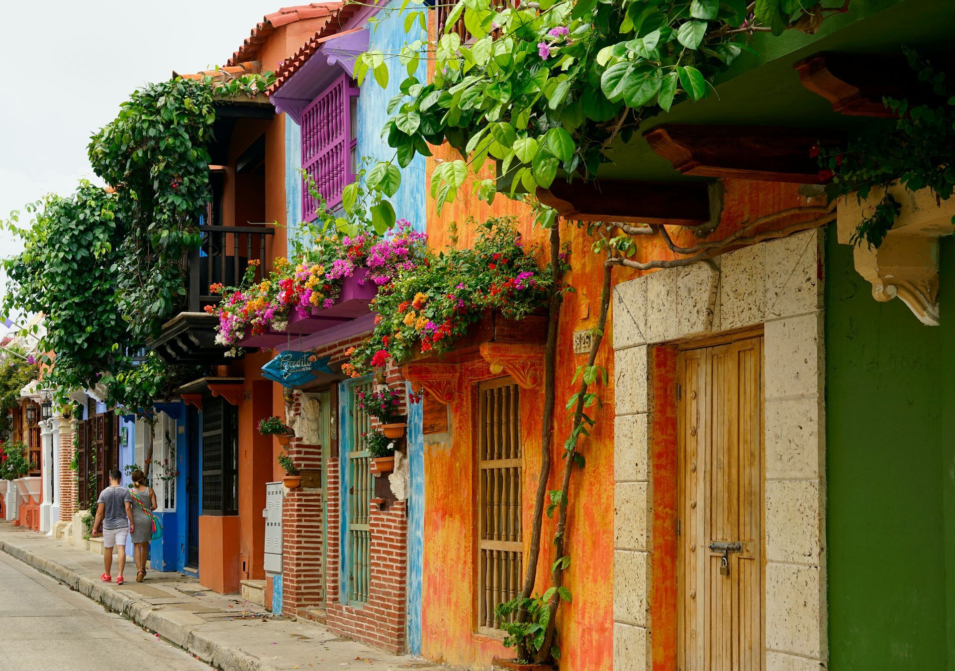 Colorful buildings with flowers and greenery line a cobblestone street. A person walks by.