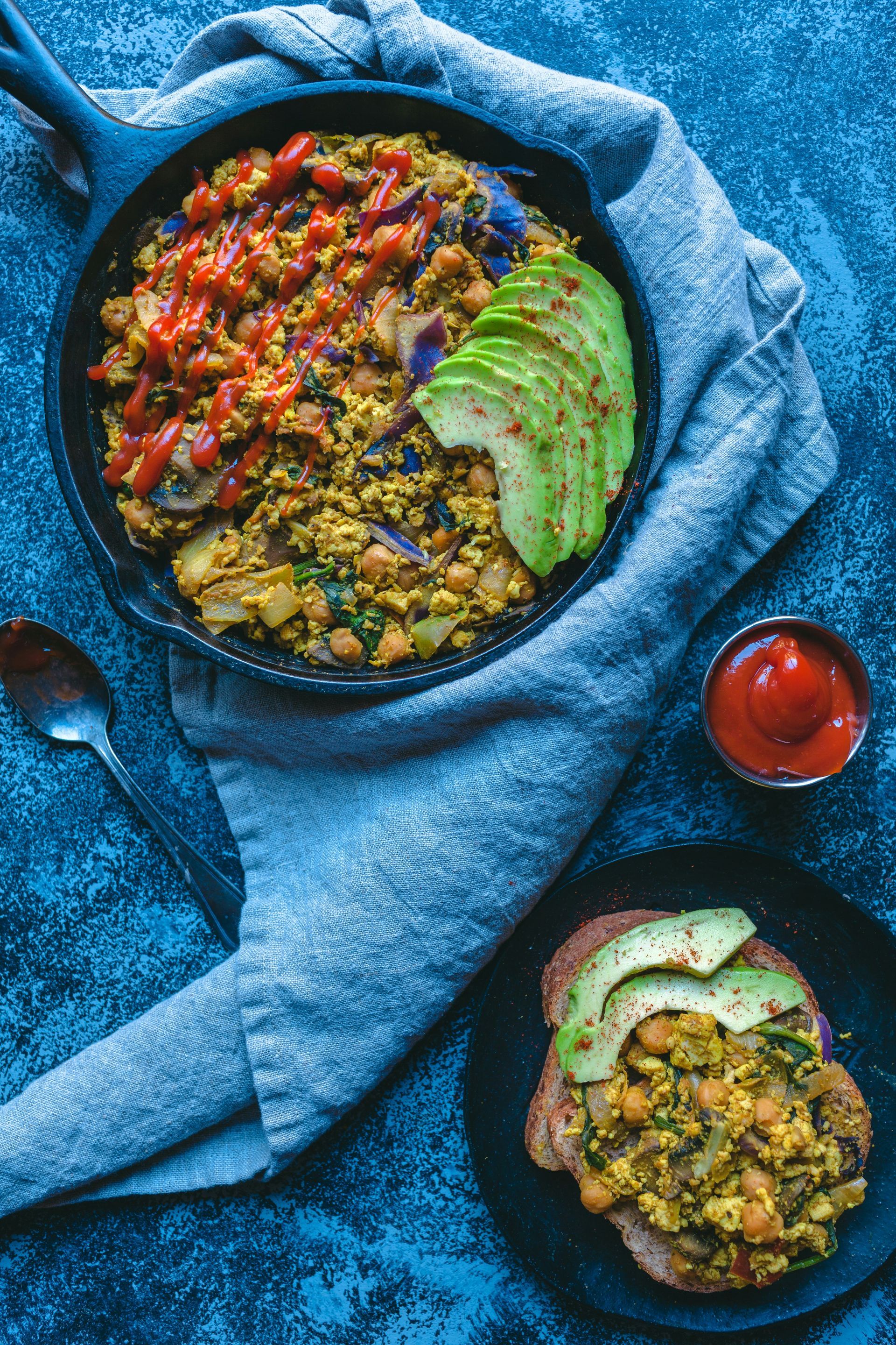 Skillet meal with tofu scramble, avocado, and tomato, served with toast and ketchup on a blue surface.
