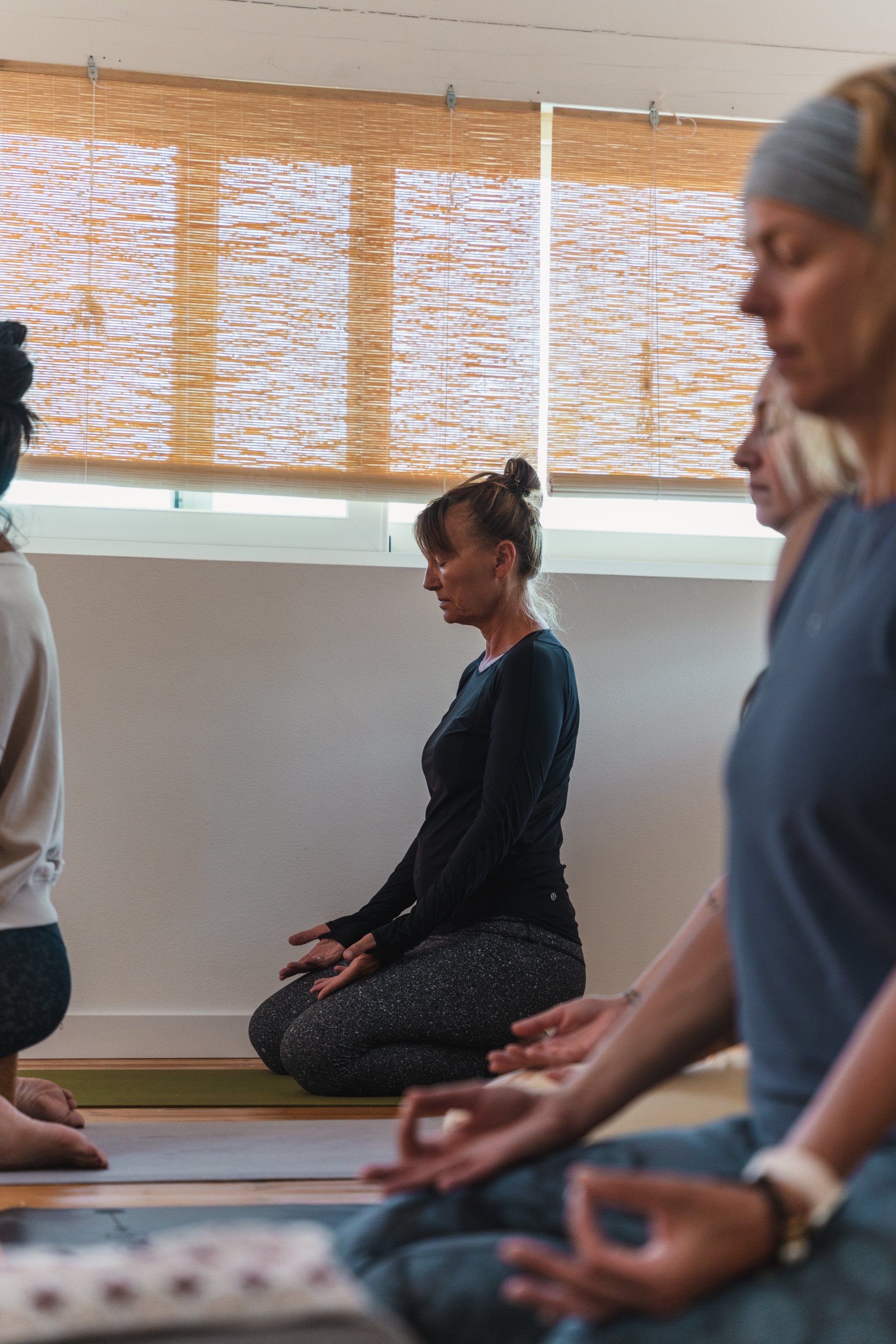 People meditating in a studio, hands in mudra, eyes closed. Natural light, wood blinds.