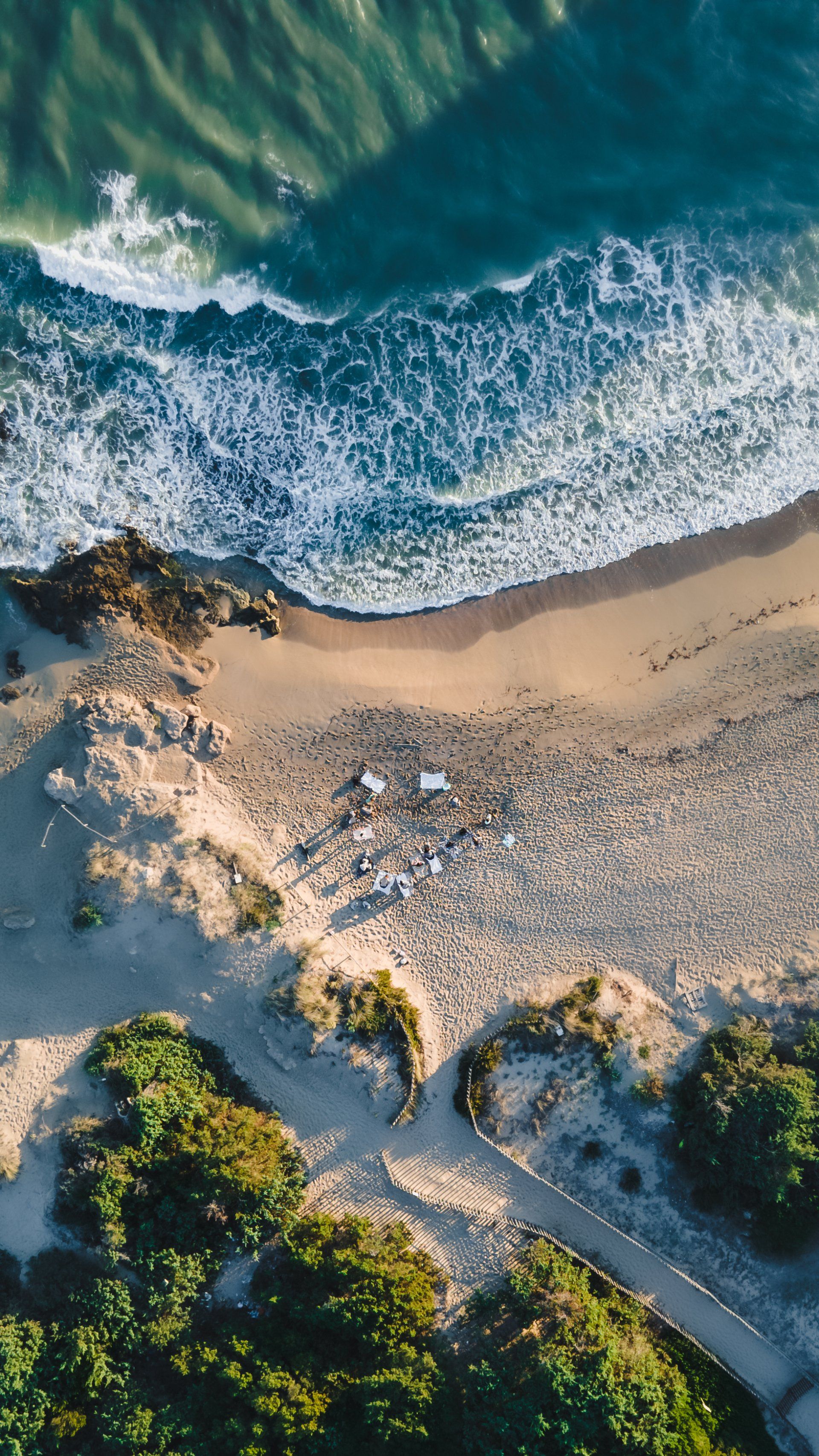 Aerial view of a beach with ocean waves, sand, dunes, and green shrubbery.