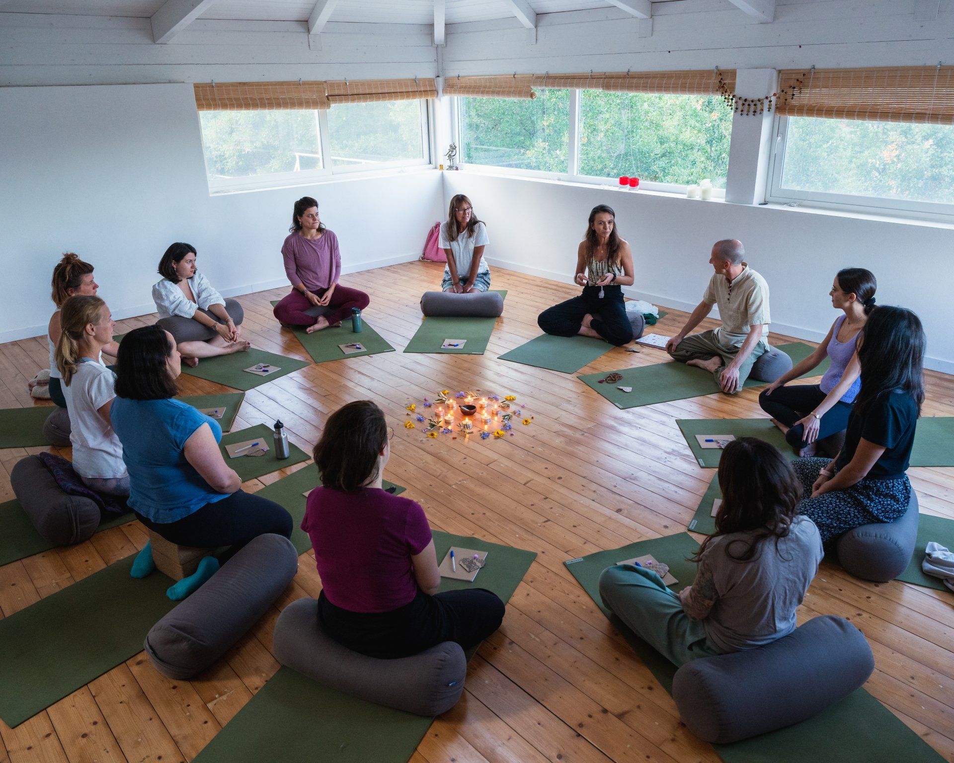 People seated in a circle on yoga mats in a light-filled room; a small centerpiece is in the middle.