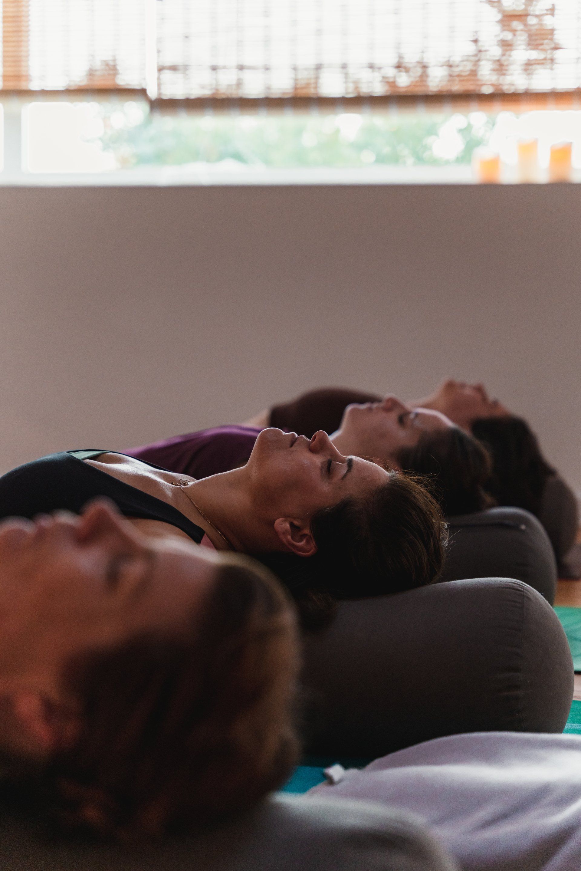 People lying back on bolsters in a yoga class, eyes closed, near a window.