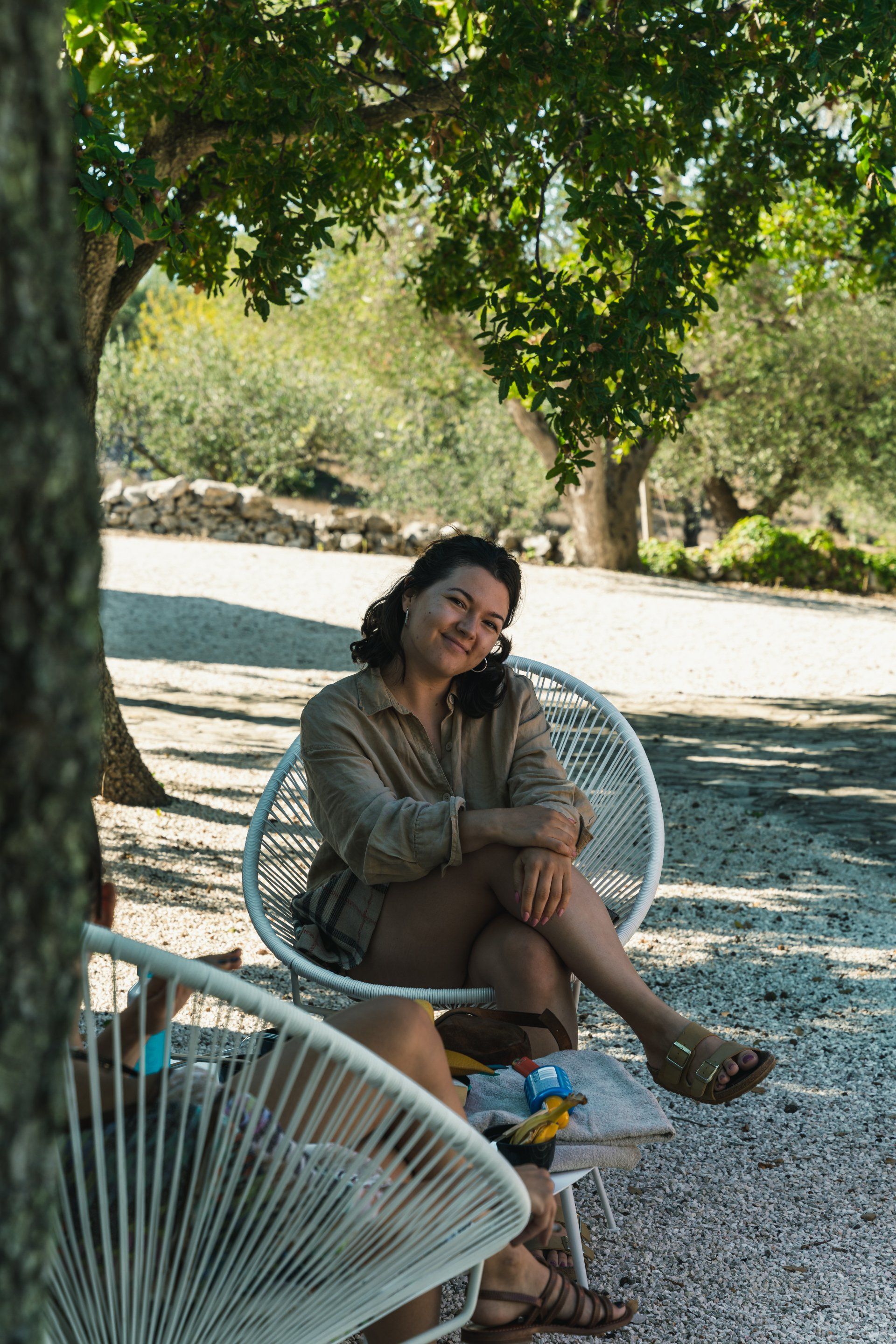 Woman in a white chair under a tree, smiling. Sunlight, natural setting, wearing a tan outfit, and sandals.