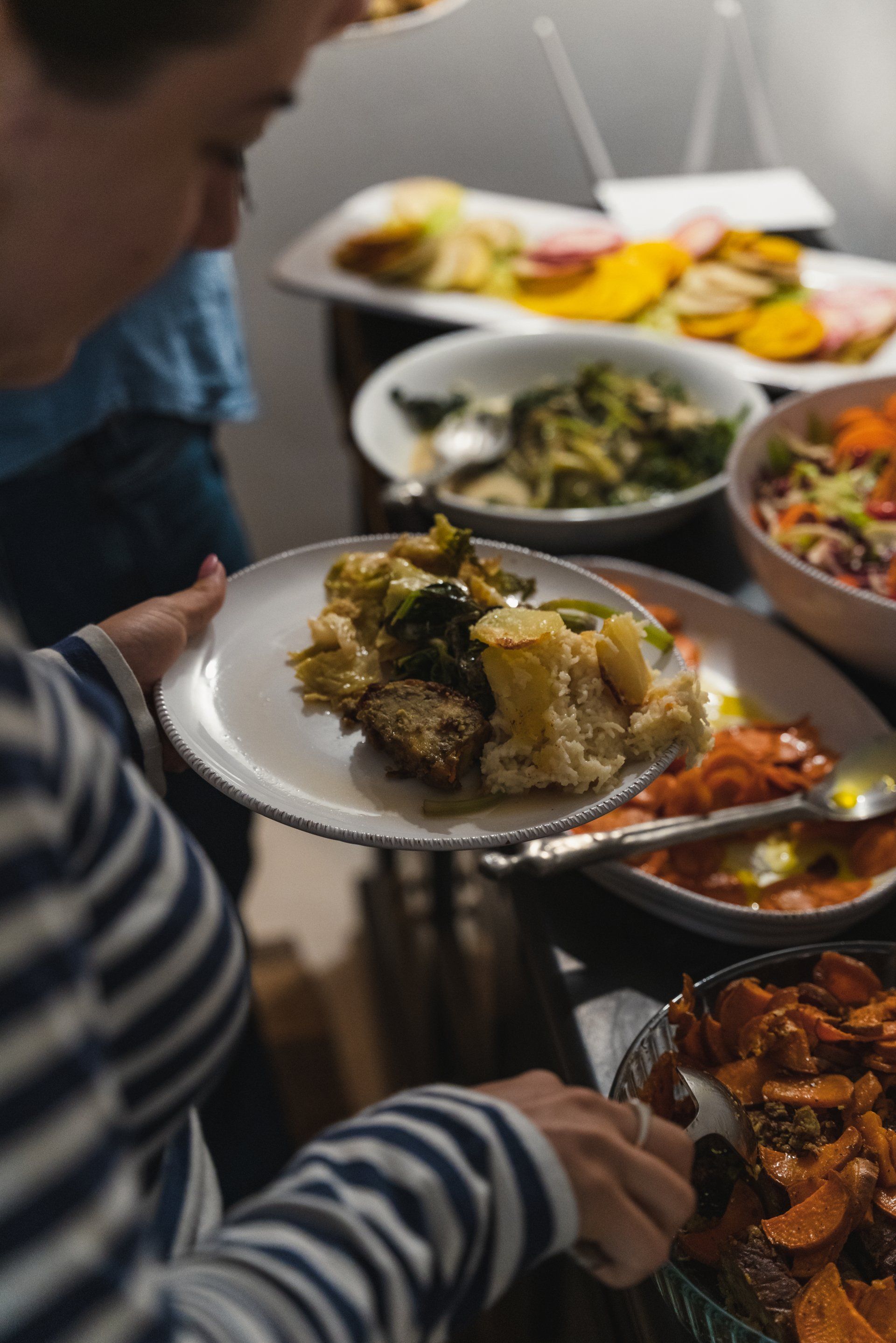 Person filling plate at a buffet table, featuring various dishes like salad and cooked vegetables.