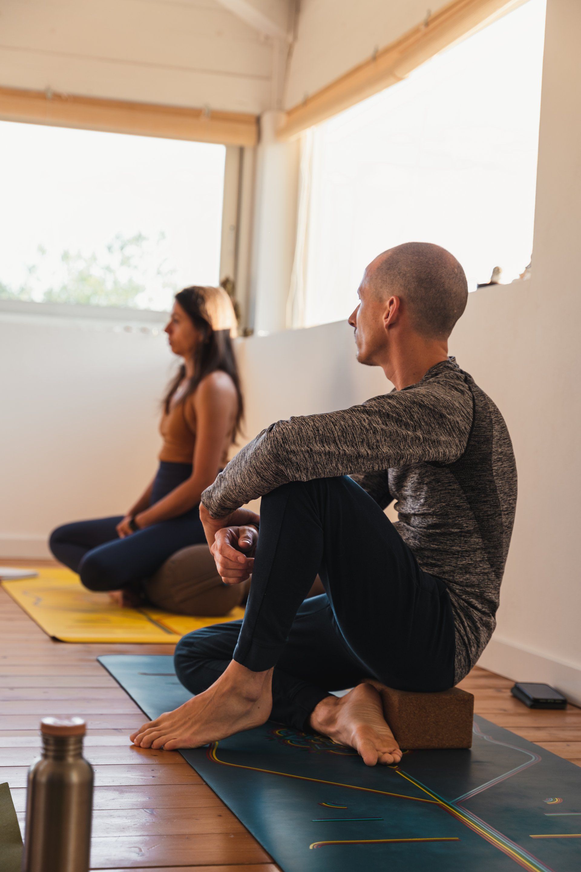 Two people meditating indoors, sitting on mats, one using a block. Natural light, peaceful setting.