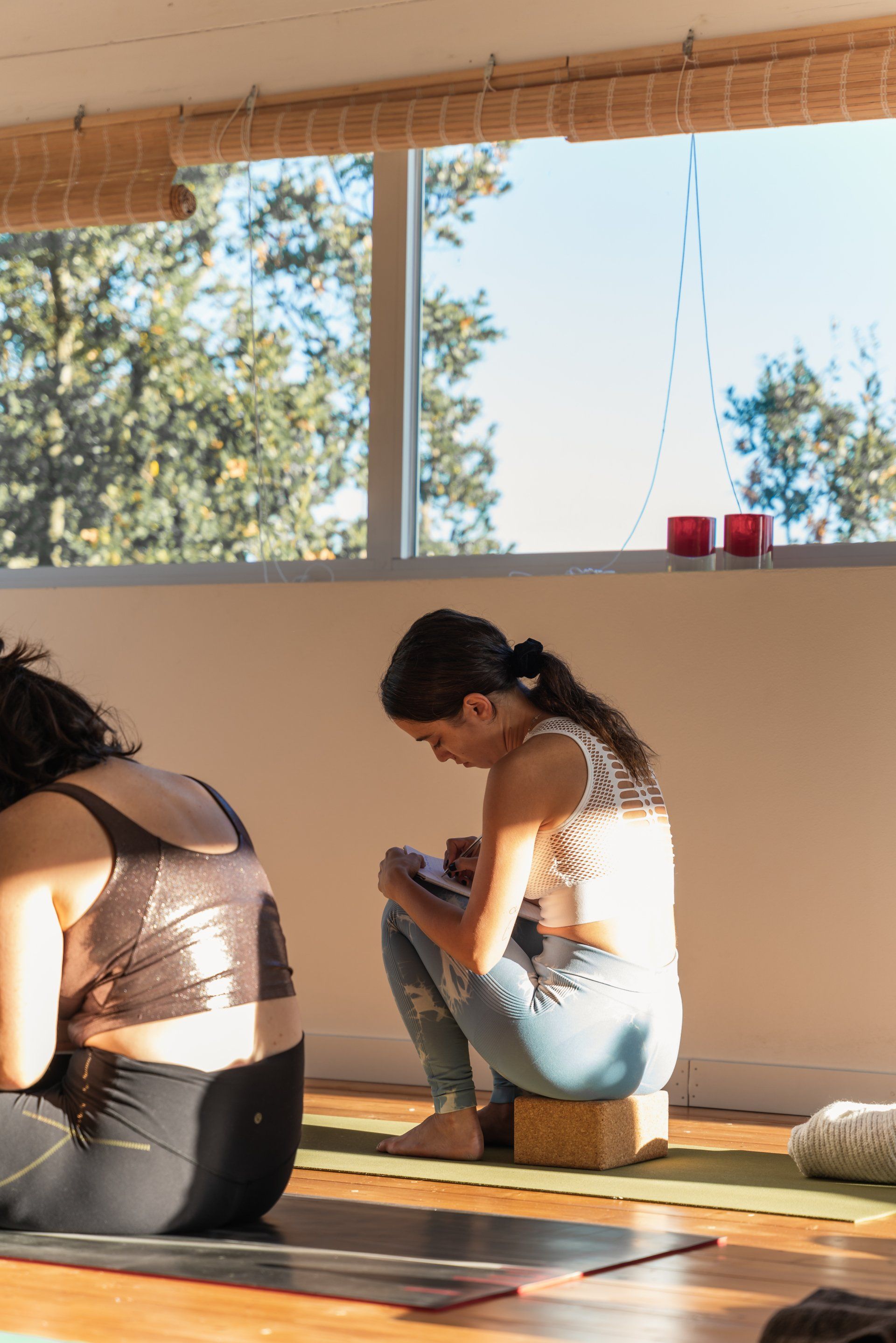 Two people seated in a yoga studio. One writes on a notepad; sunlight streams in the window.