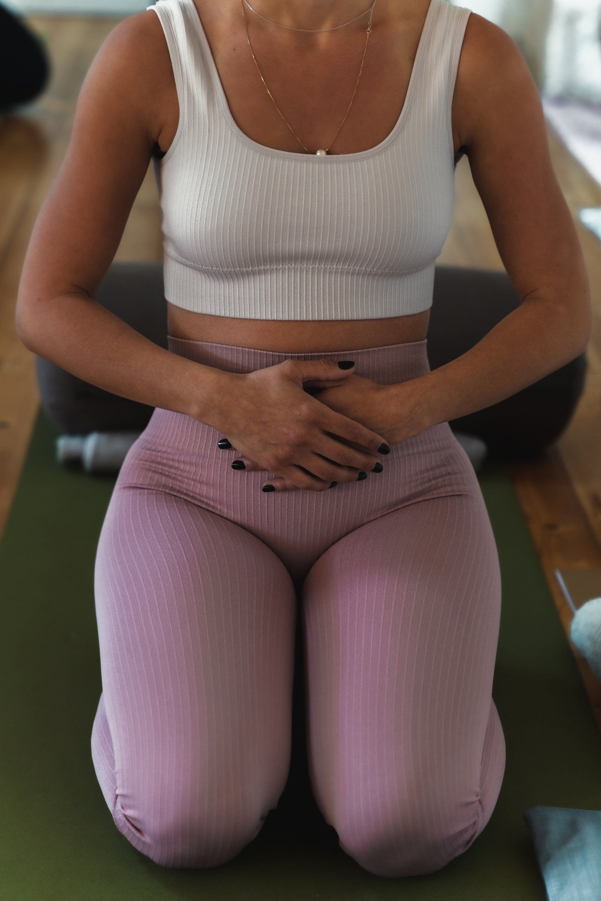 Woman in workout clothes meditating, hands on stomach, kneeling on a mat. Beige top and pink leggings.