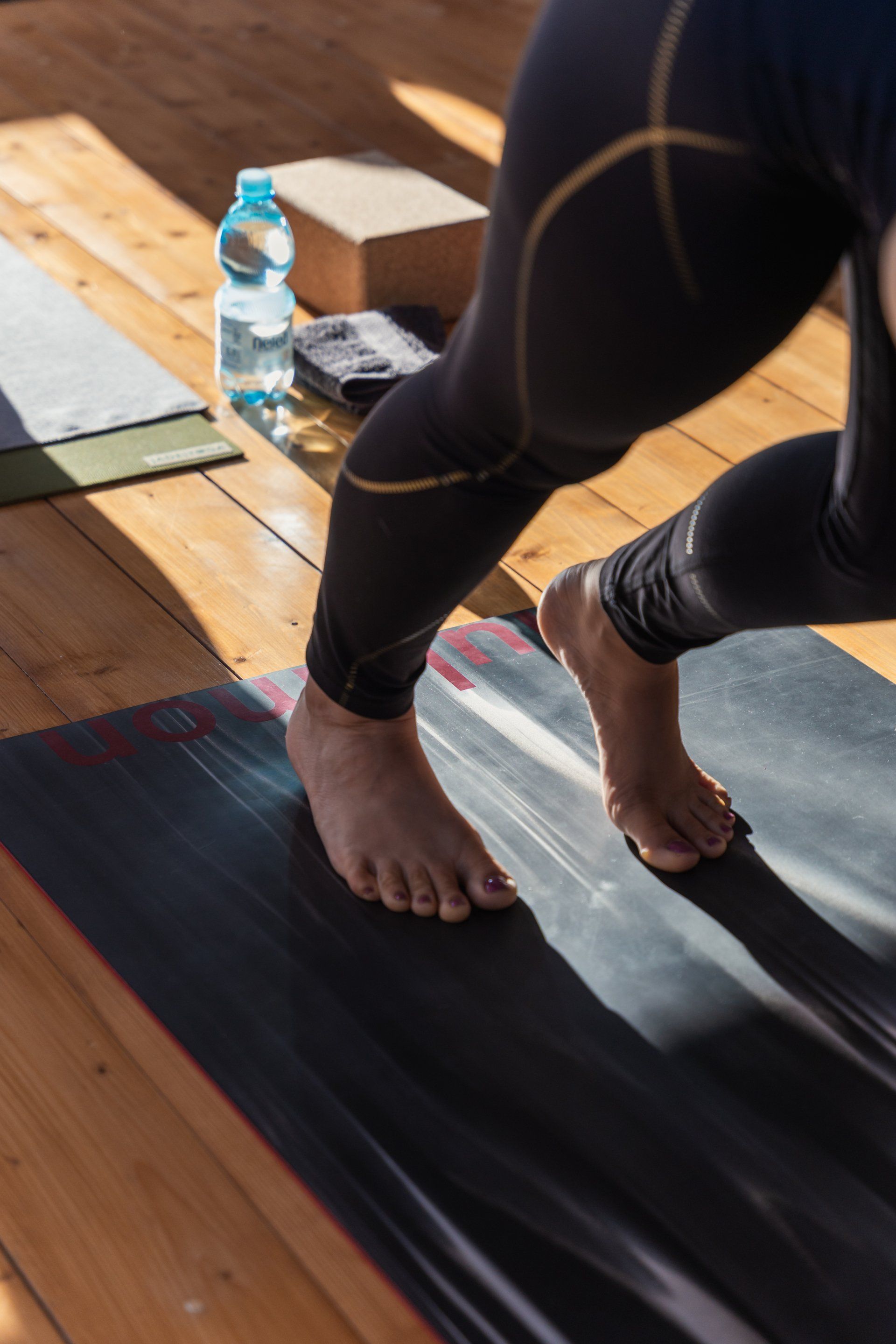 Person in black leggings on yoga mat, about to step. Wooden floor, water bottle, and block visible.