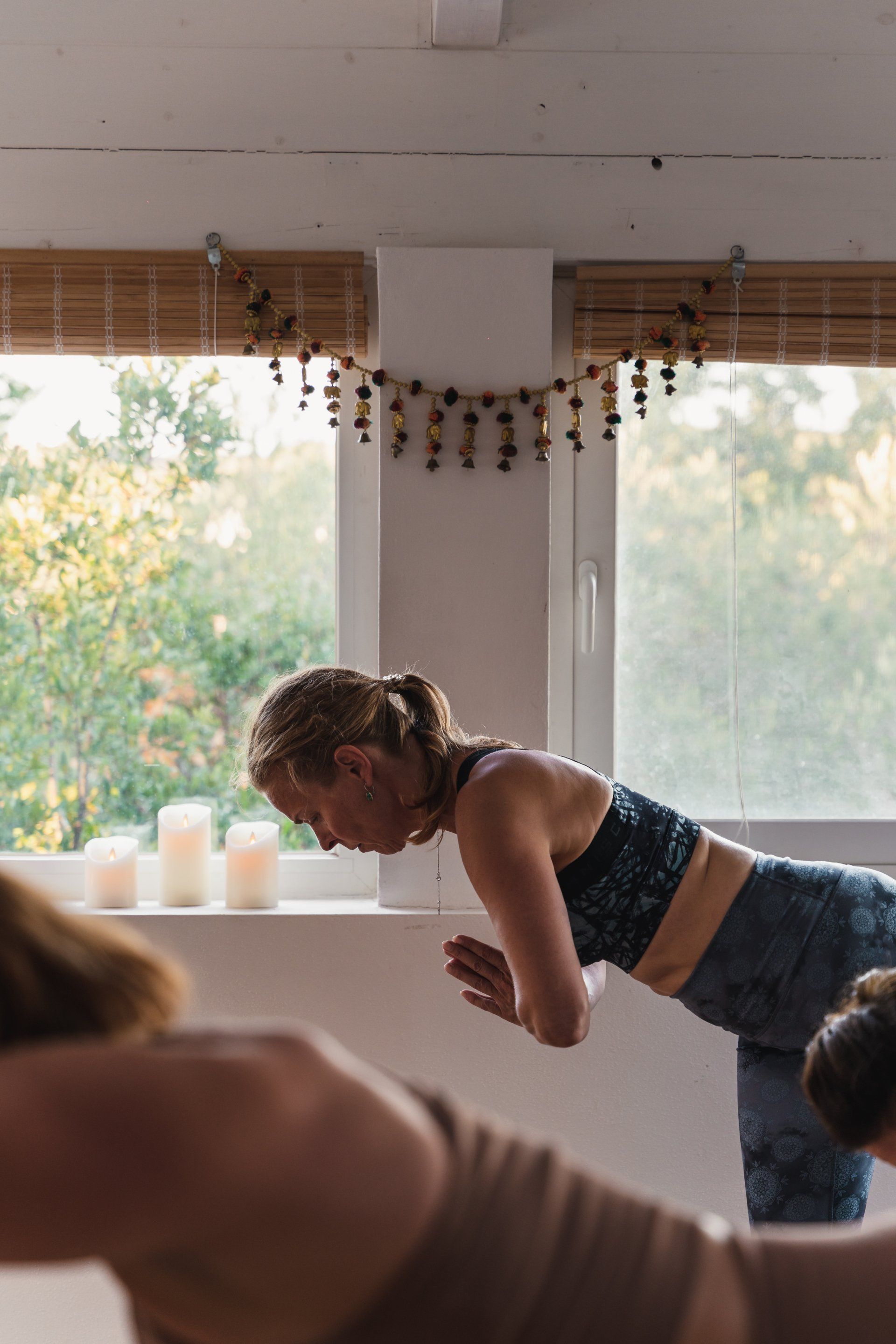 Woman in yoga pose, hands together, in a bright room with a window and candles.