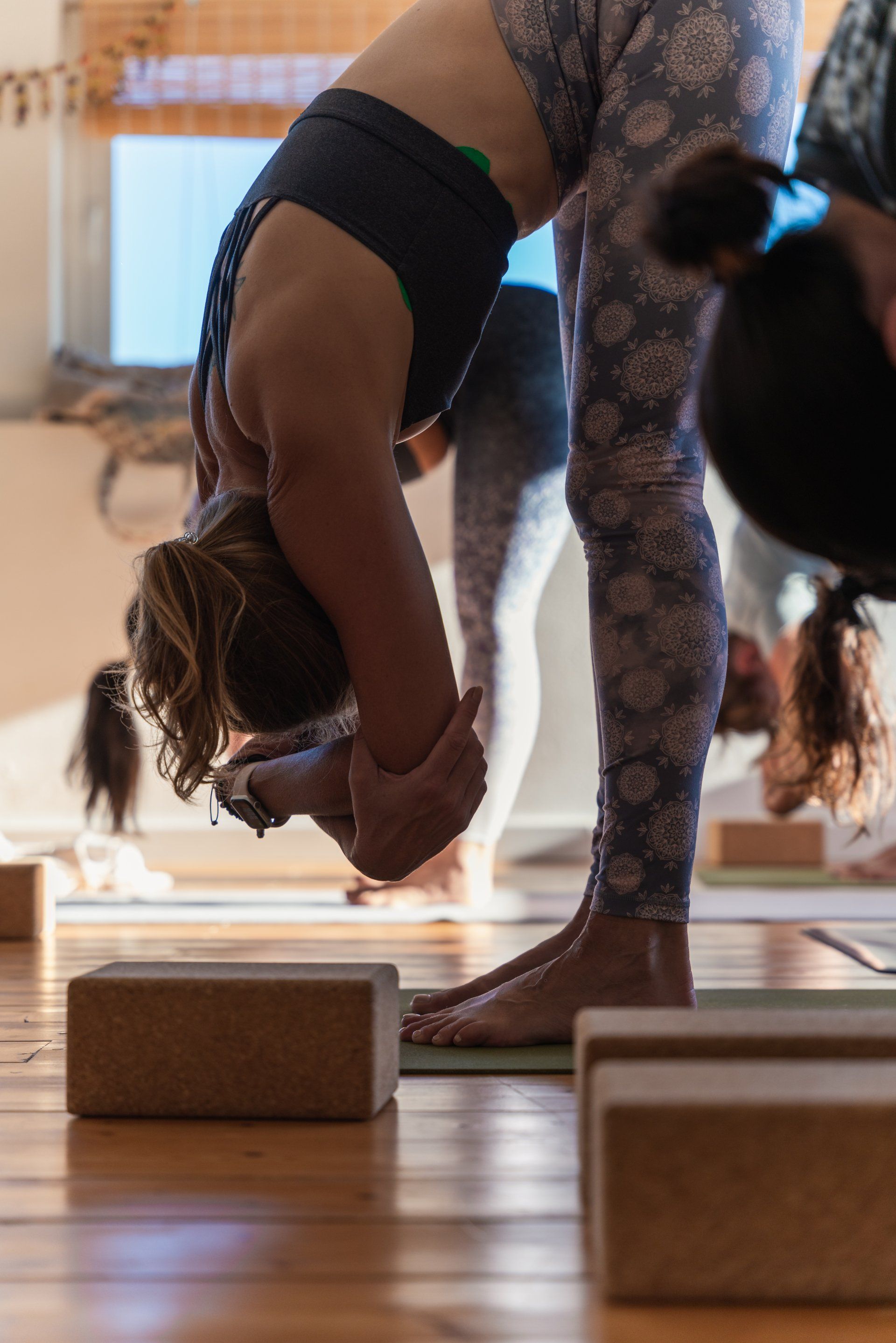 Woman in yoga class, bent forward, hands on feet, floral leggings, wooden floor.
