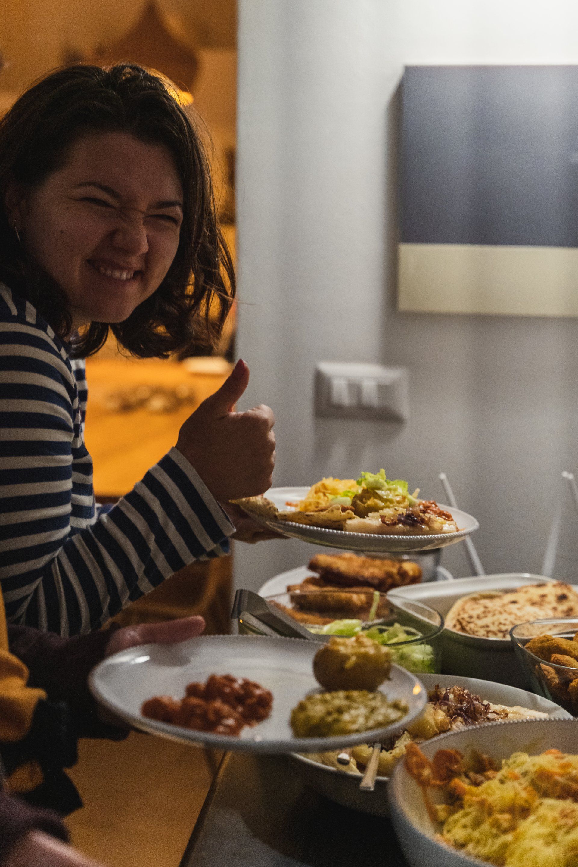 Woman with striped shirt smiles, giving a thumbs-up while holding plates of food at a buffet.