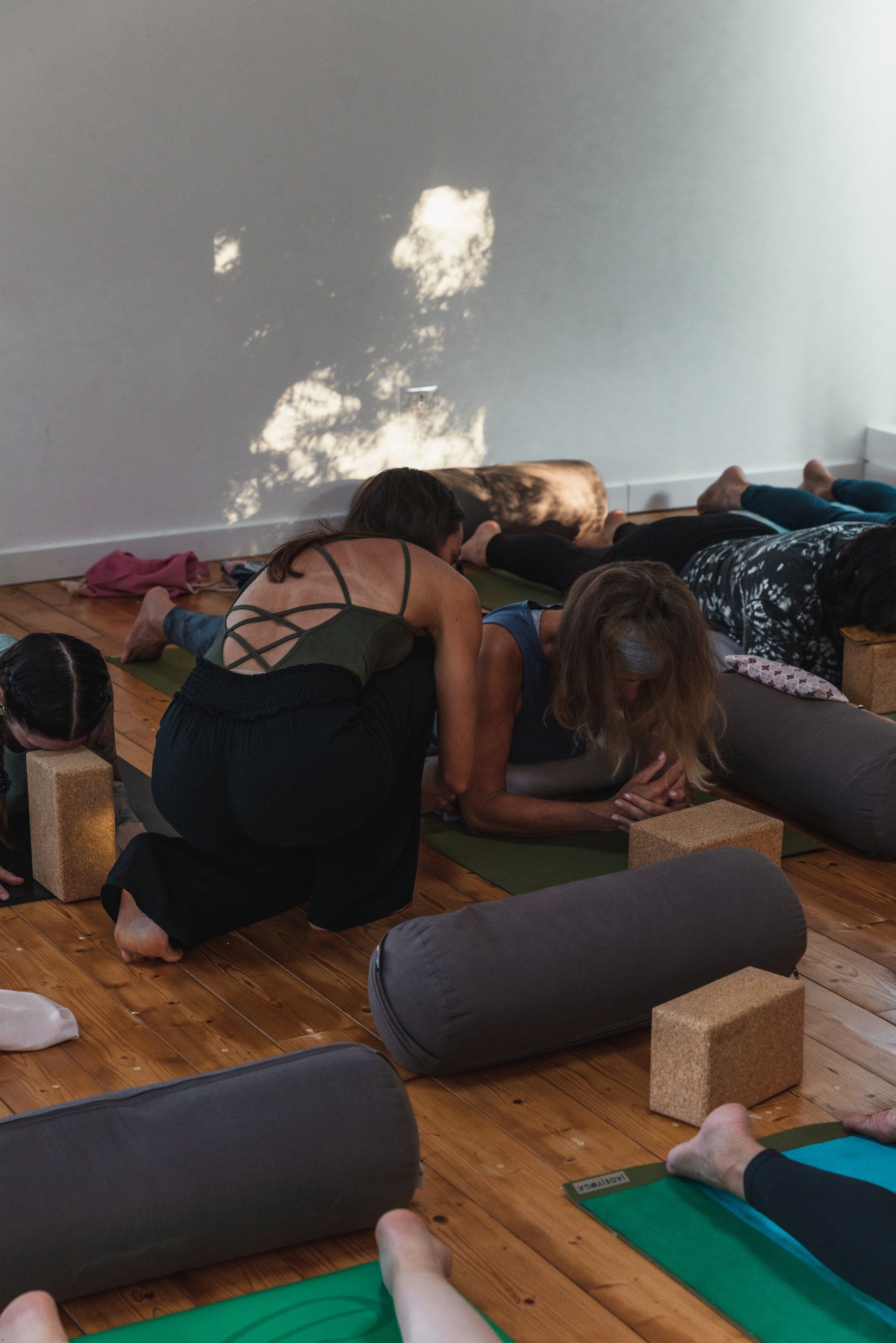 Yoga instructor adjusting a person's pose in a studio. Others rest on mats with bolsters.