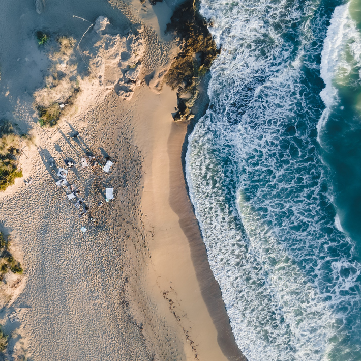 Sandy beach meets ocean with foamy waves, small group of people with white tables and chairs.