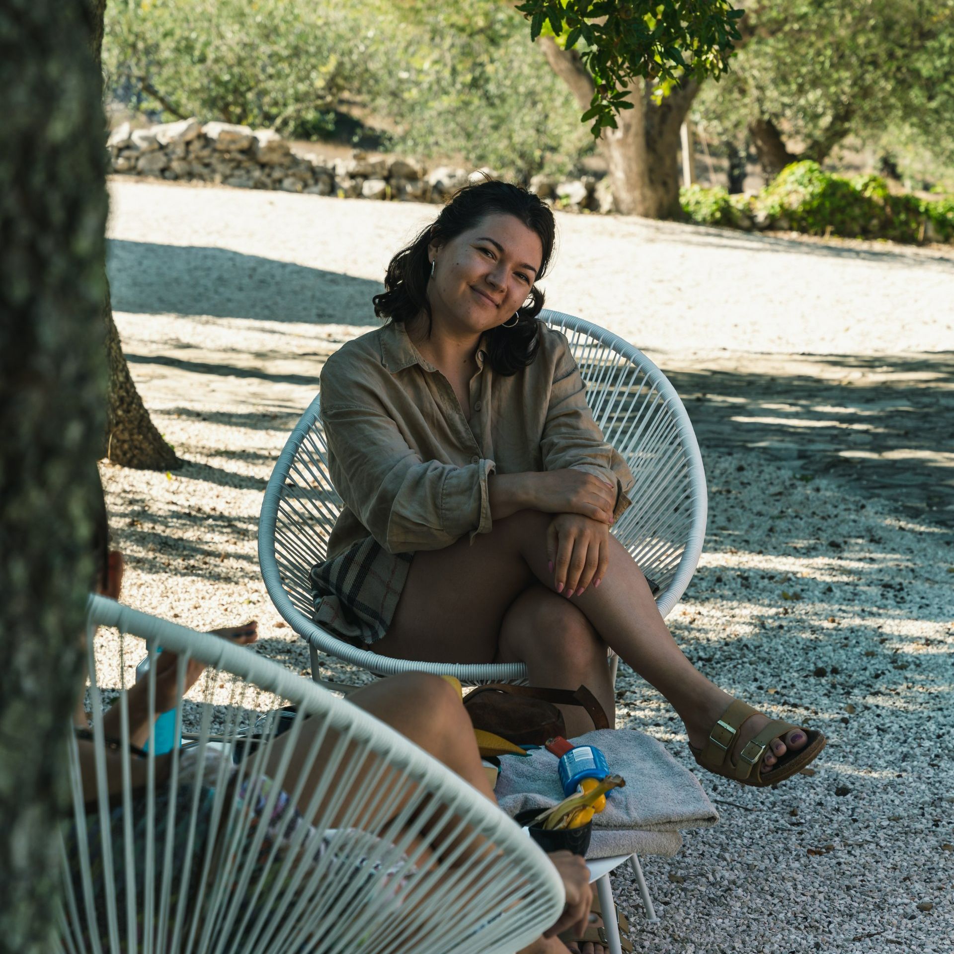 Woman sitting in white wicker chair outdoors, smiling, wearing tan shirt and sandals.
