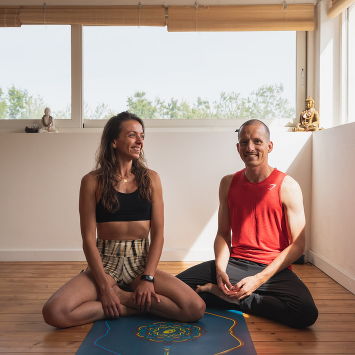Woman and man seated on yoga mats, smiling, in a bright room with window.