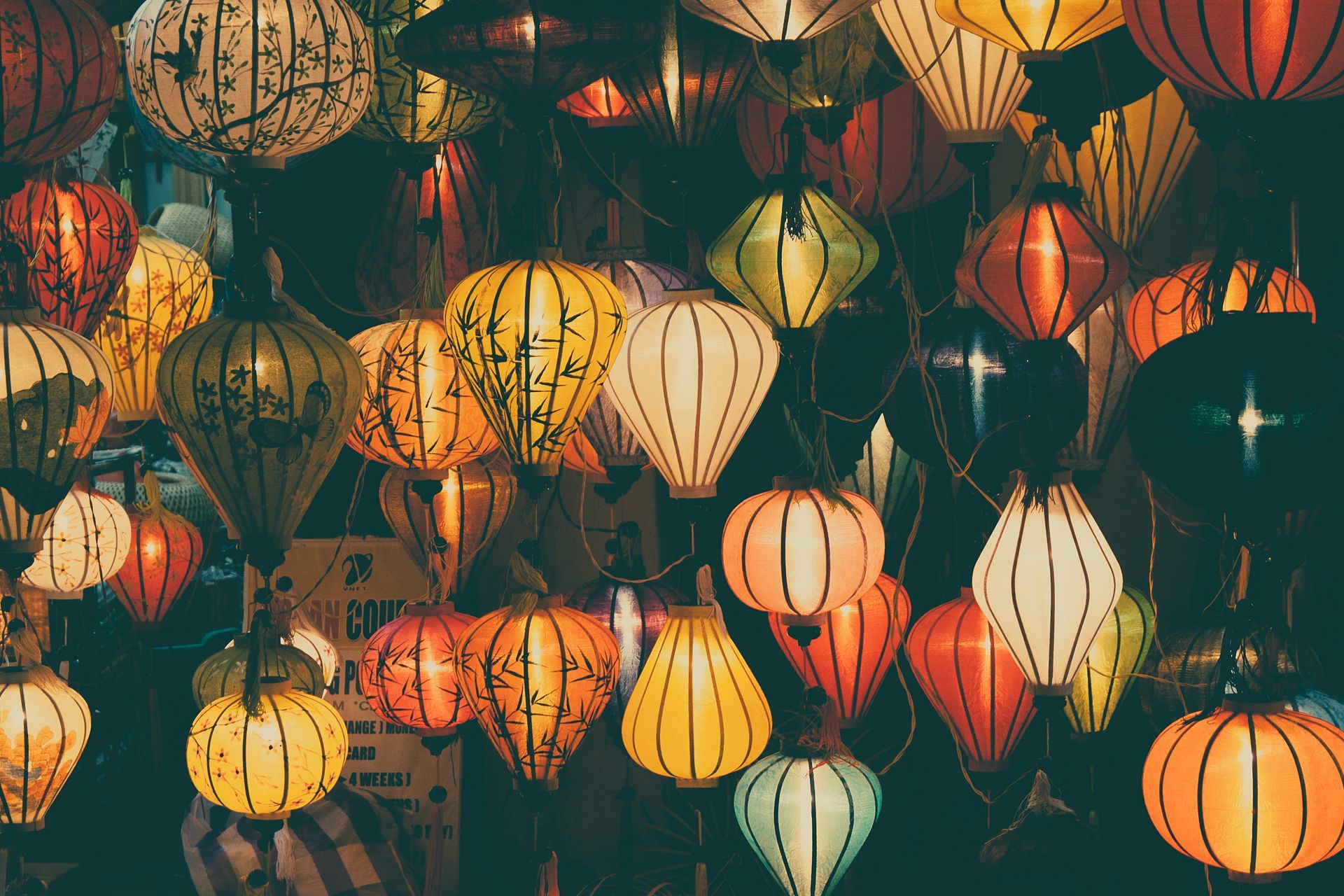 Colorful paper lanterns hanging, illuminated. Yellow, orange, and white lanterns in a dark setting.