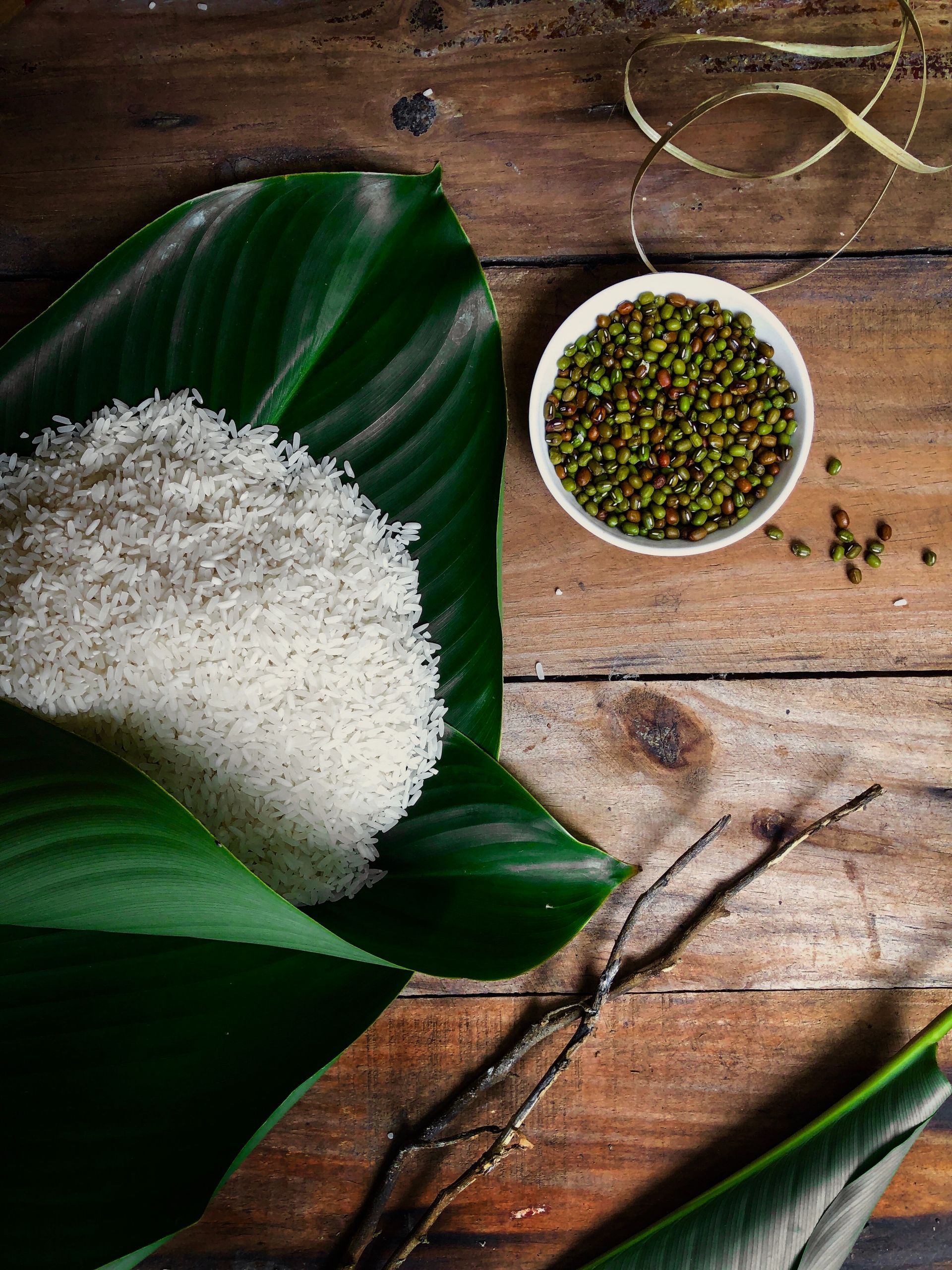 Rice pile and bowl of green mung beans on wooden surface, with green leaves.