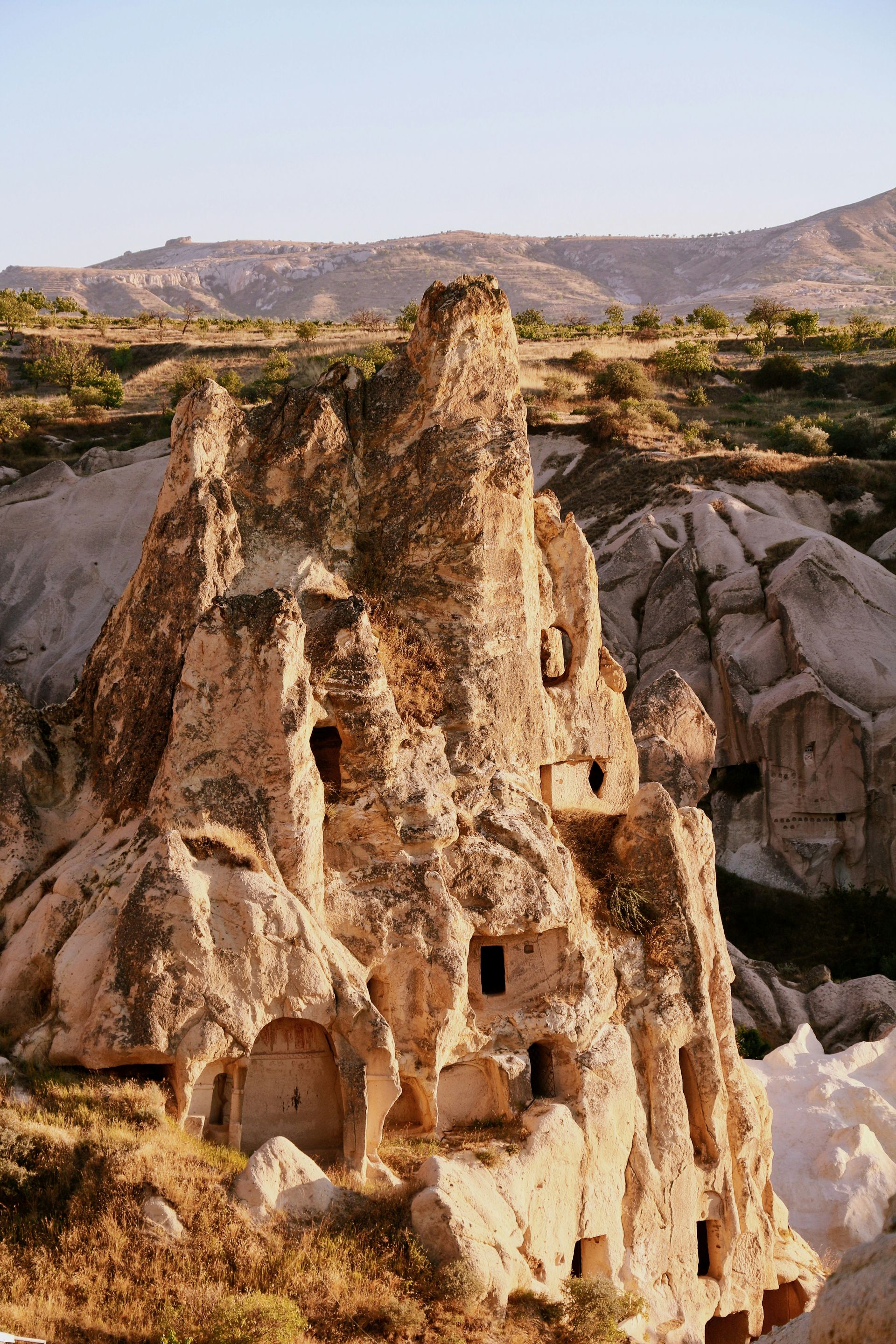 Rock formations with carved-out dwellings in a mountainous region, beige and brown hues.