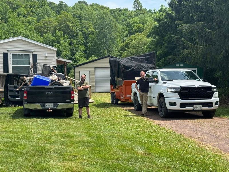 Two men stand near a white truck and a black truck with a trailer, parked on grass in front of a house.