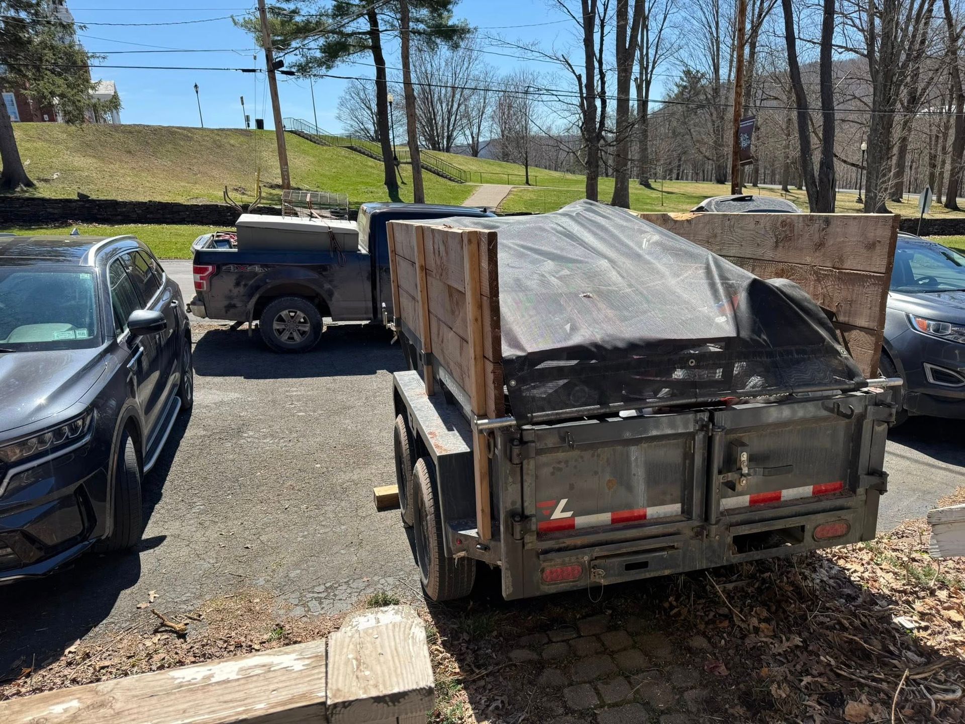 Pickup truck loaded with a covered dumpster trailer in a parking lot near trees and a lake