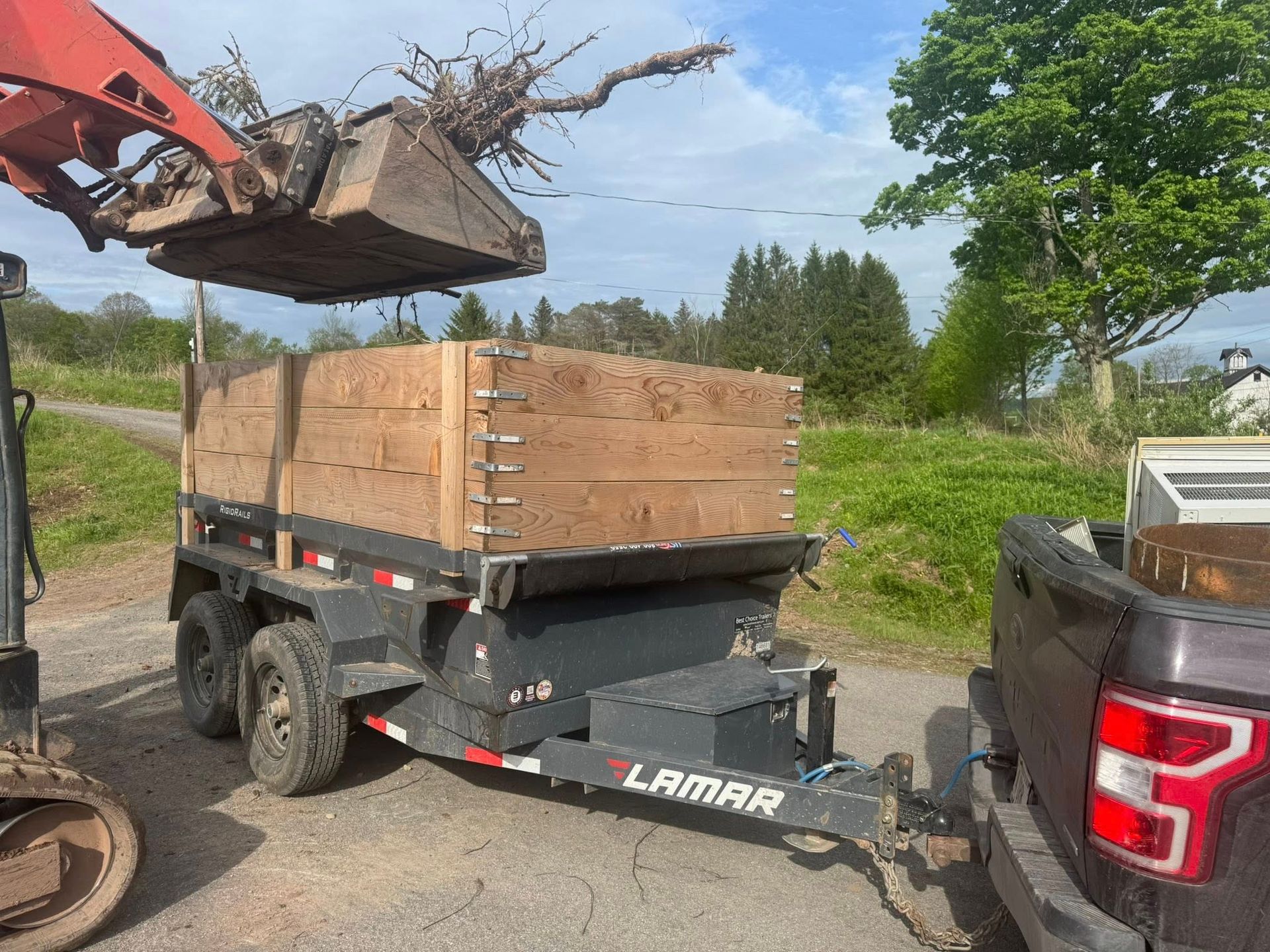 Excavator loading wooden crates onto a black utility trailer in a grassy outdoor area
